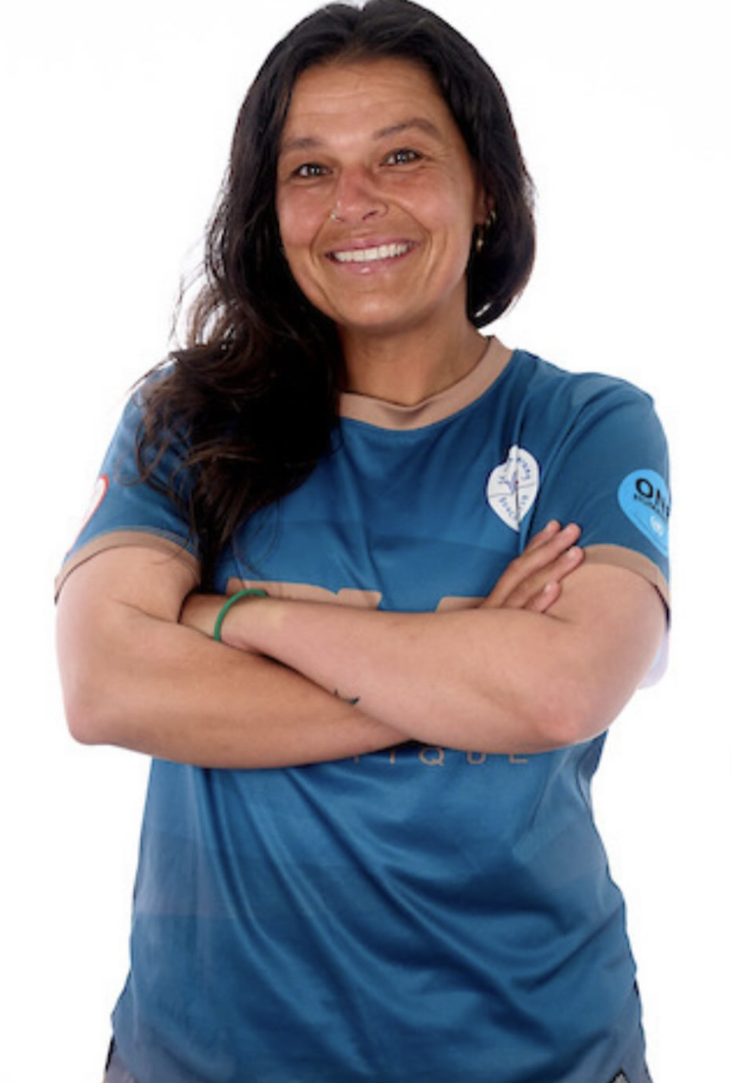 A woman with long dark hair smiling and crossing her arms. She is wearing a blue sports shirt with logos on the sleeves.
