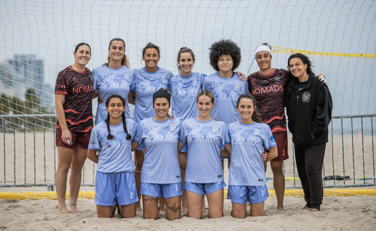 Group of female beach volleyball players posing on sand court, some wearing blue jerseys with 'NOMADS' logo, others in maroon and black jerseys, with a volleyball net in the background.