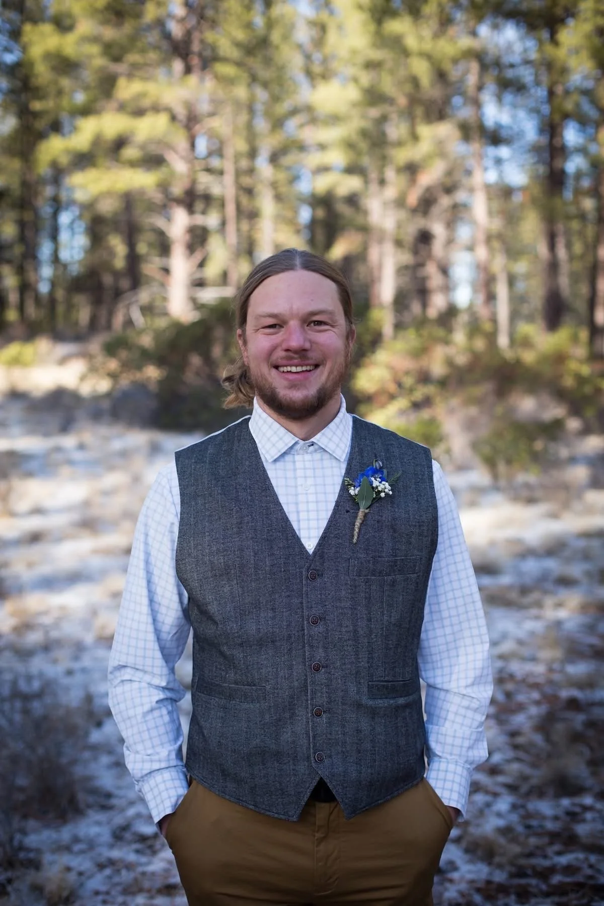 A smiling man with long hair and a beard, dressed in a white checkered shirt, a dark vest with a boutonniere, and brown pants, standing outdoors in a forested area with trees and a rocky, snowy ground.