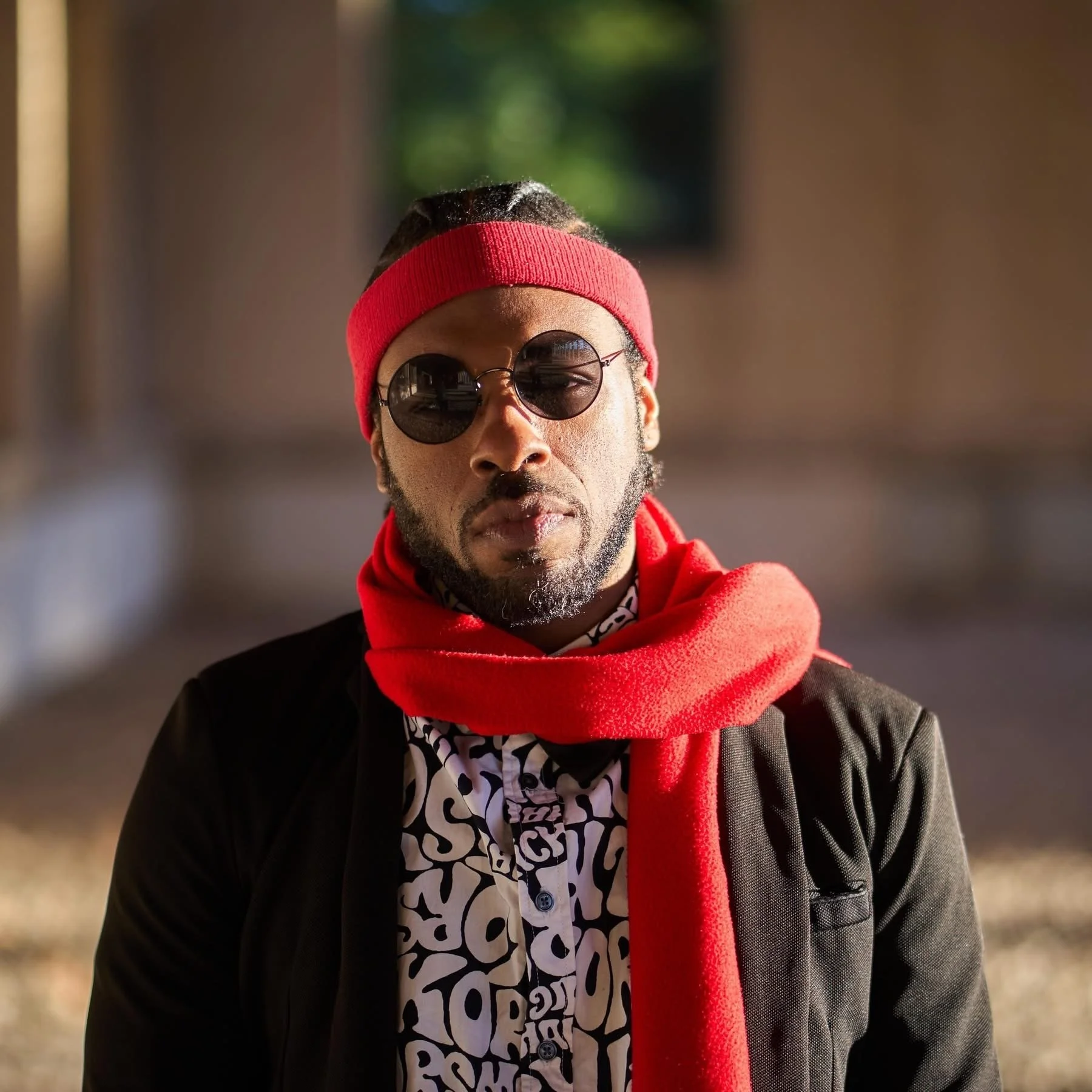 A man with a beard and dark skin wearing round sunglasses, a red headband, a red scarf, a black jacket, and a patterned shirt underneath, standing outdoors with blurred background.