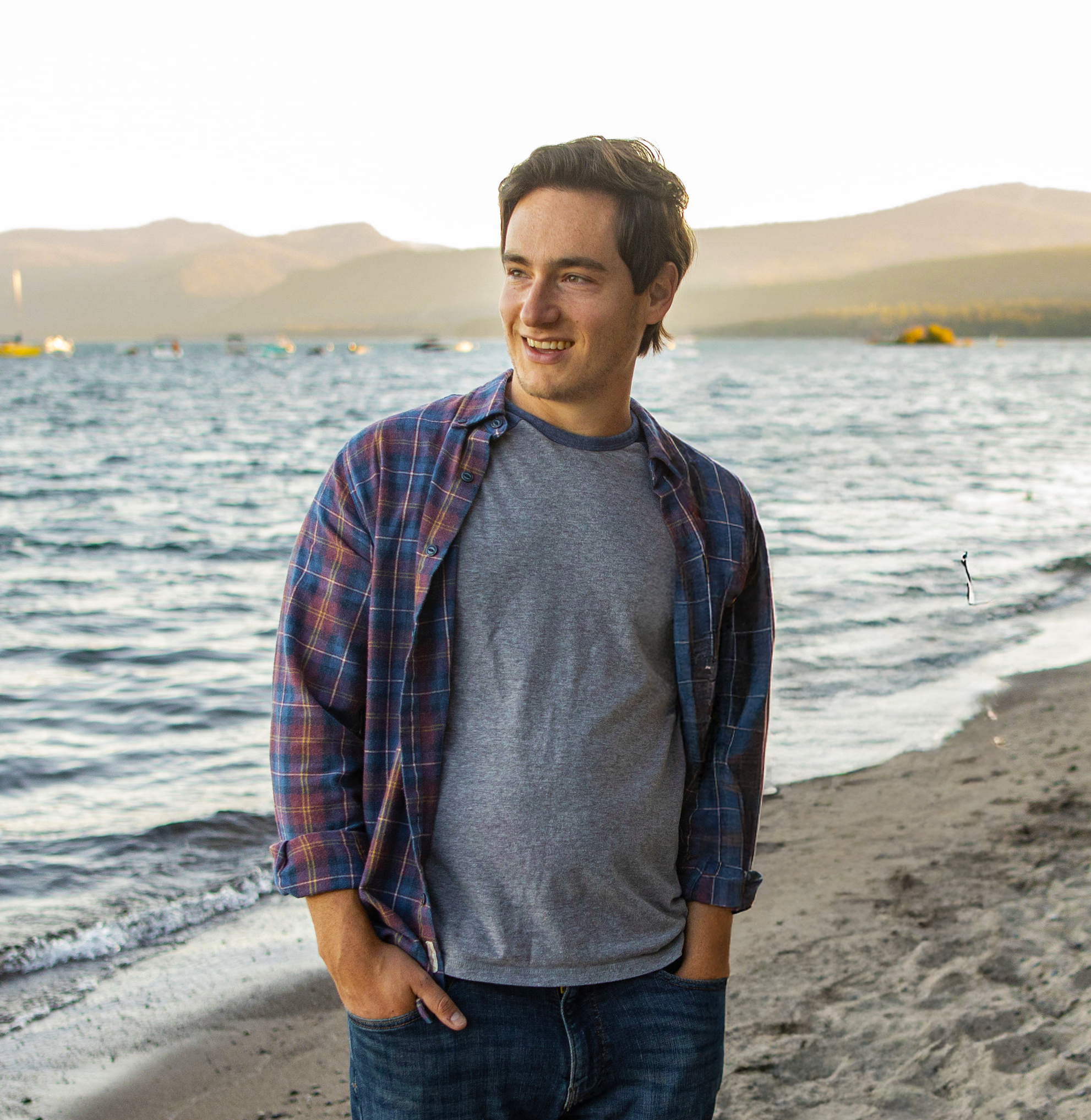 A young man with dark hair, wearing a grey t-shirt and a blue plaid shirt, stands near a body of water with boats and mountains in the background, smiling and looking to the side.