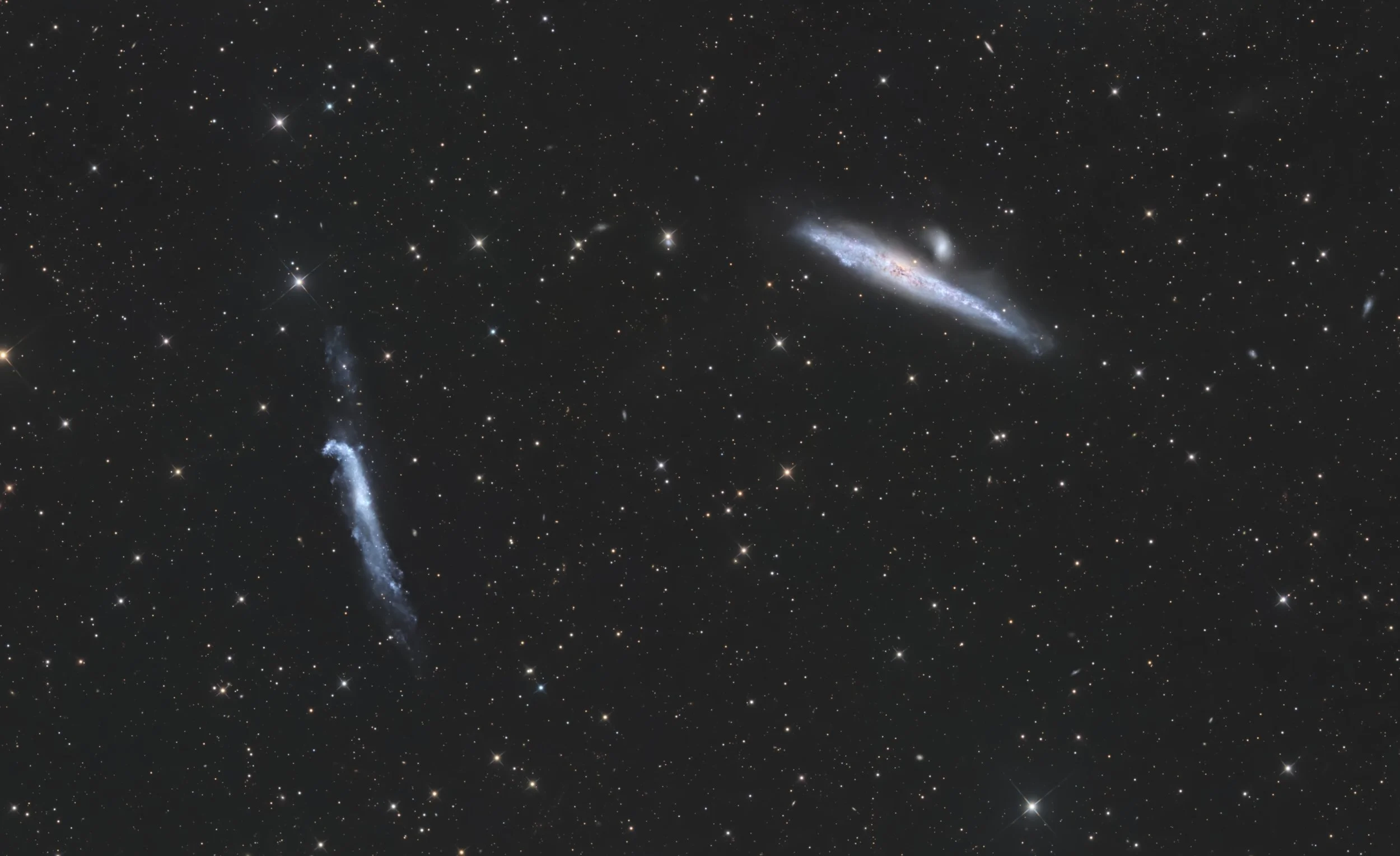 A panoramic view of deep space showing numerous stars, galaxies, and nebulae, with a few prominent galaxies featuring bright cores and elongated shapes.