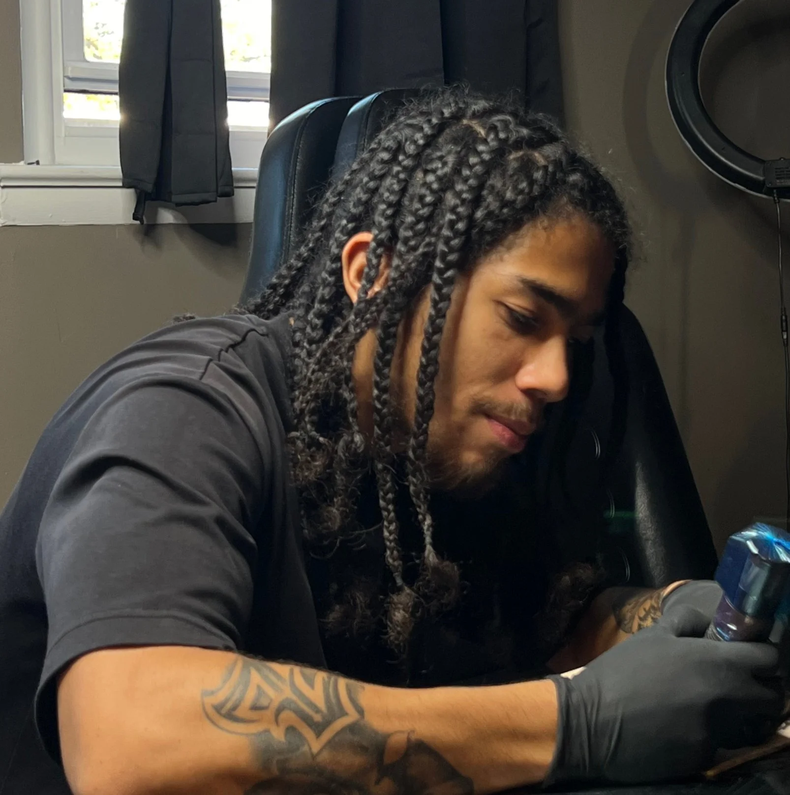 A man with braided hair, wearing a black shirt, sitting indoors near a window, looking down, with a tattoo on his right arm, and a water bottle in front of him.