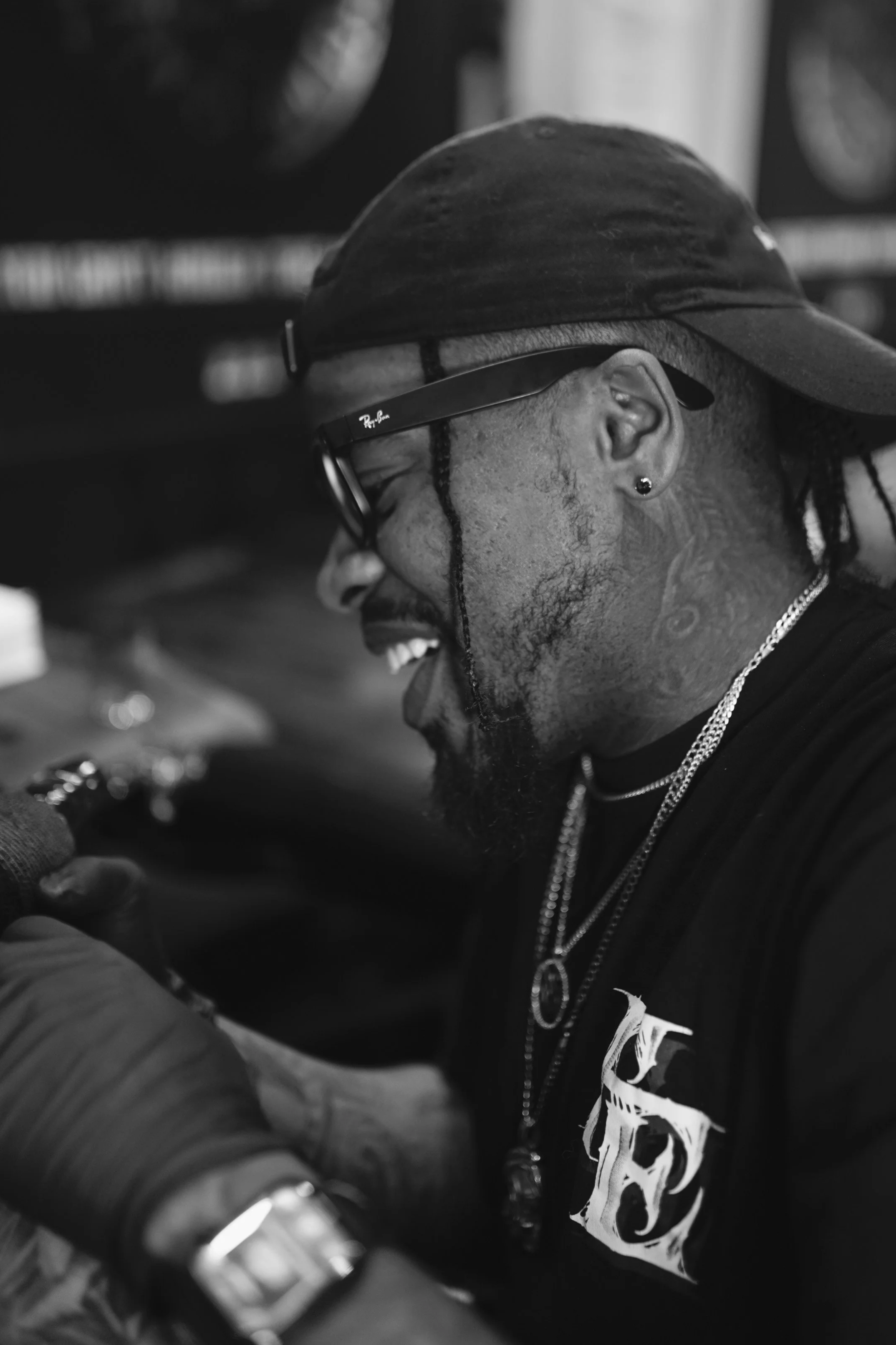 Close-up black and white photo of a man wearing a cap, glasses, earrings, and jewelry, smiling and looking down.