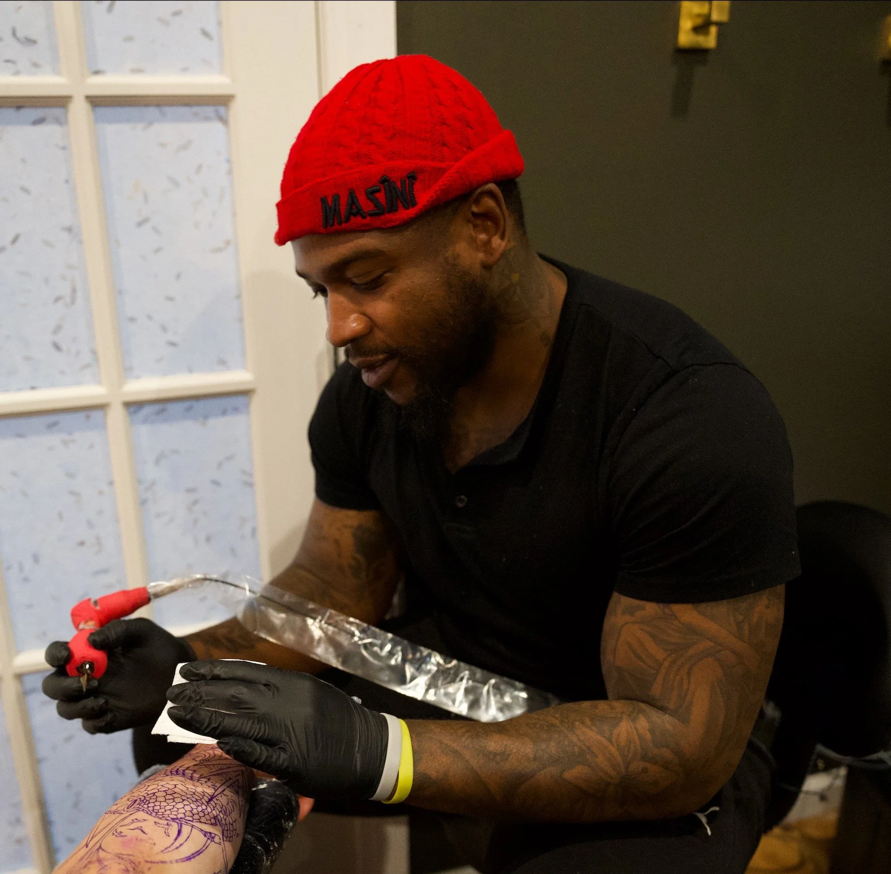 A man with a red knit beanie with the word 'MASSIV' embroidered on it, tattooed arms, and wearing black gloves, is working on a tattoo machine, tattooing a person's arm. The scene is indoors with a door in the background.