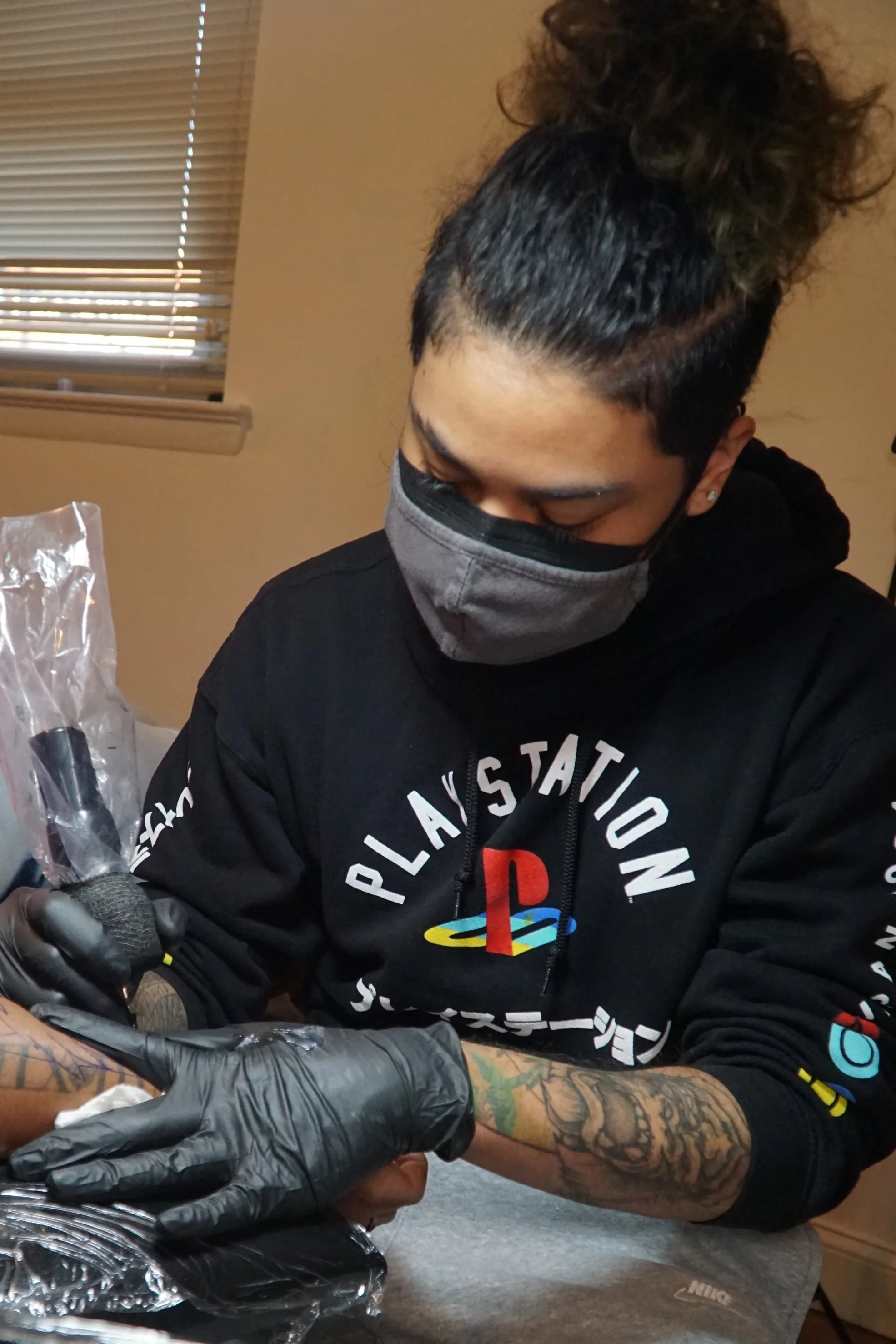 A person with tattoos on their arm and wearing a black hoodie with a PlayStation logo on it is getting a tattoo. The tattoo artist is wearing black gloves and is working on the person's forearm at a table with tattoo equipment, in a room with beige walls and a window with blinds.