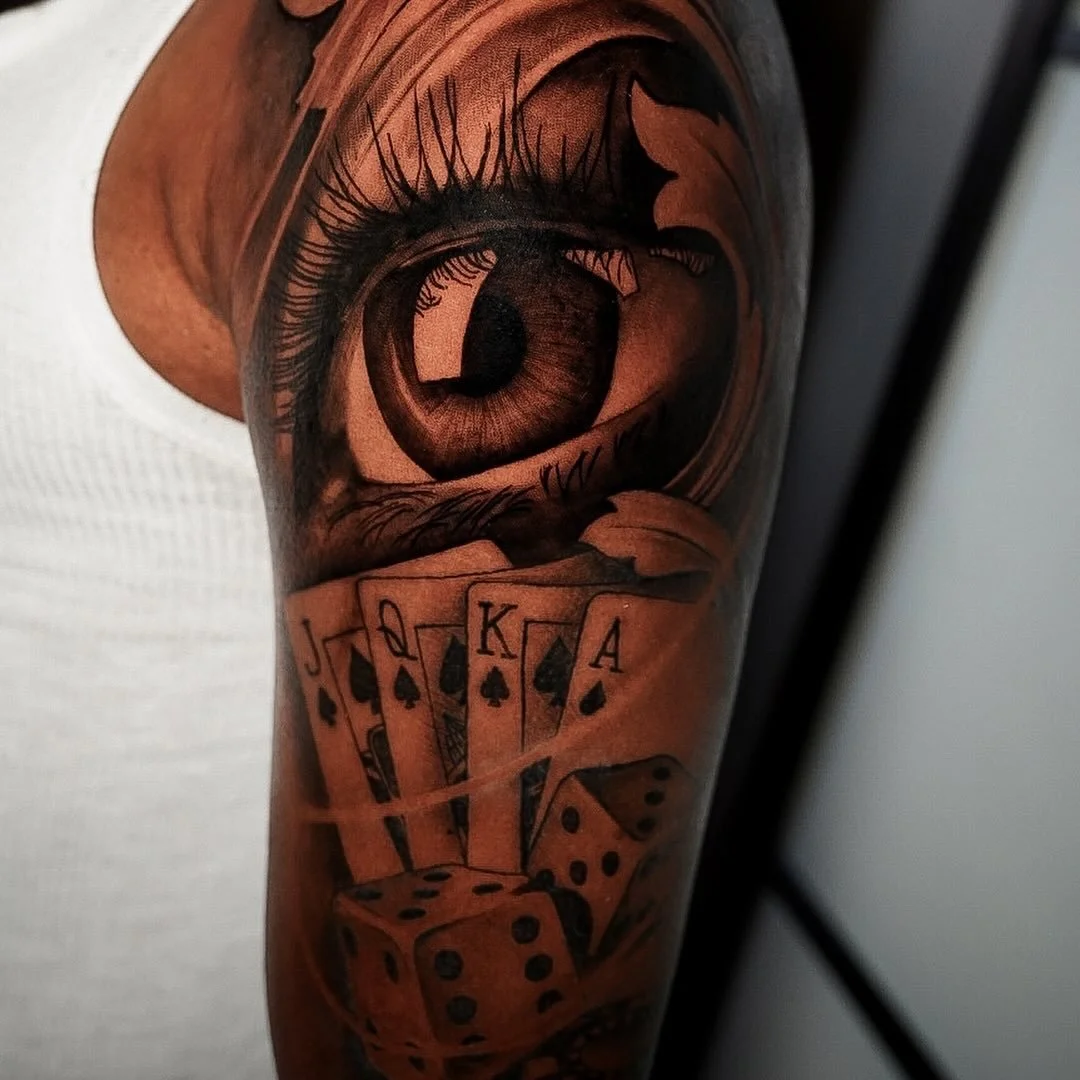 Tattoo of a realistic eye with detailed eyelashes and iris, above a set of playing cards (J, Q, K, A of spades) and dice.