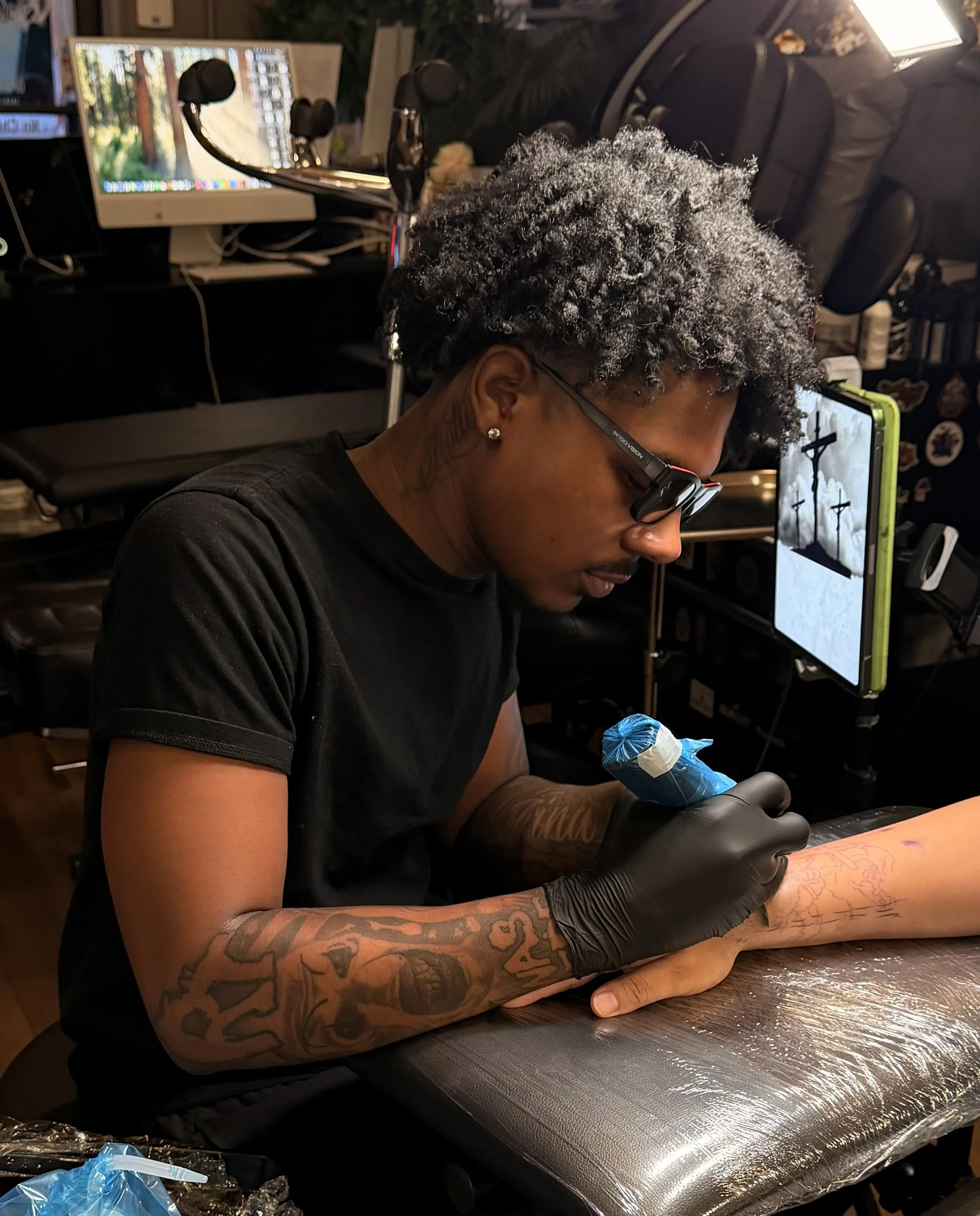 A person with gray dreadlocks, wearing glasses and black gloves, is sitting at a table and creating a tattoo on a client's forearm using a tattoo machine. The background shows a computer monitor, headphones, and tattoo supplies in a tattoo studio.