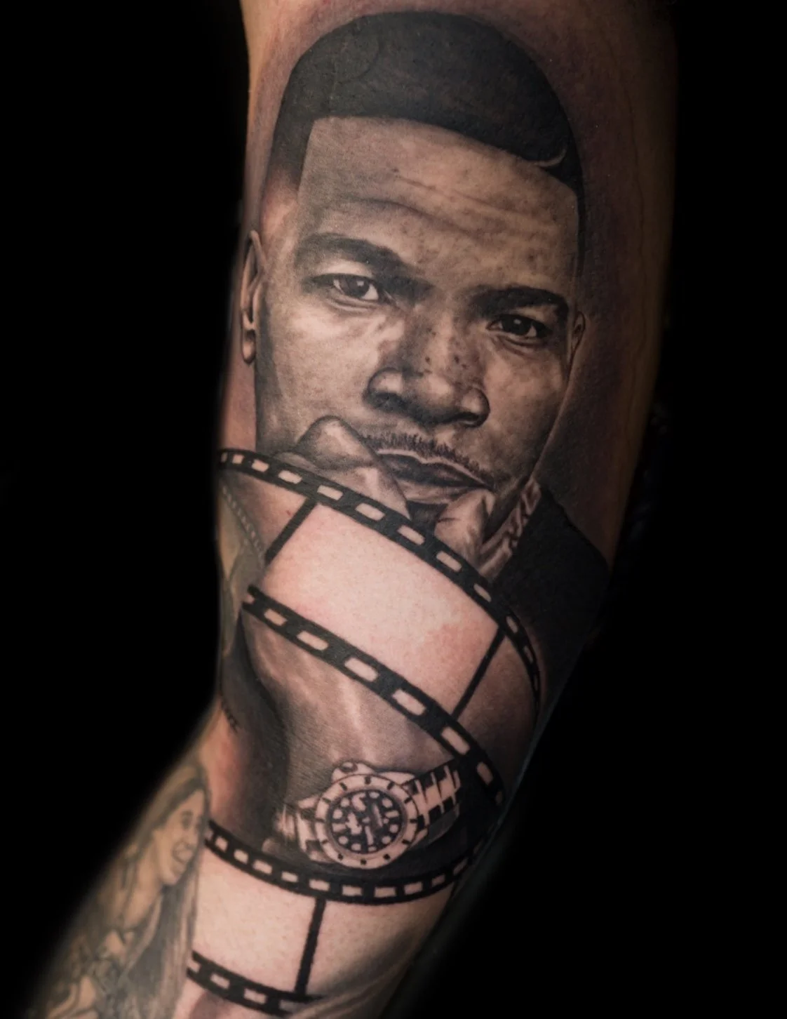 Black and gray tattoo of a young man's face with a chain around his neck on a person's arm.