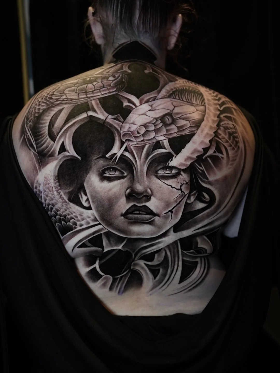 Black and white tattoo on a person's back depicting a woman's face surrounded by snakes, with cracks on her face.