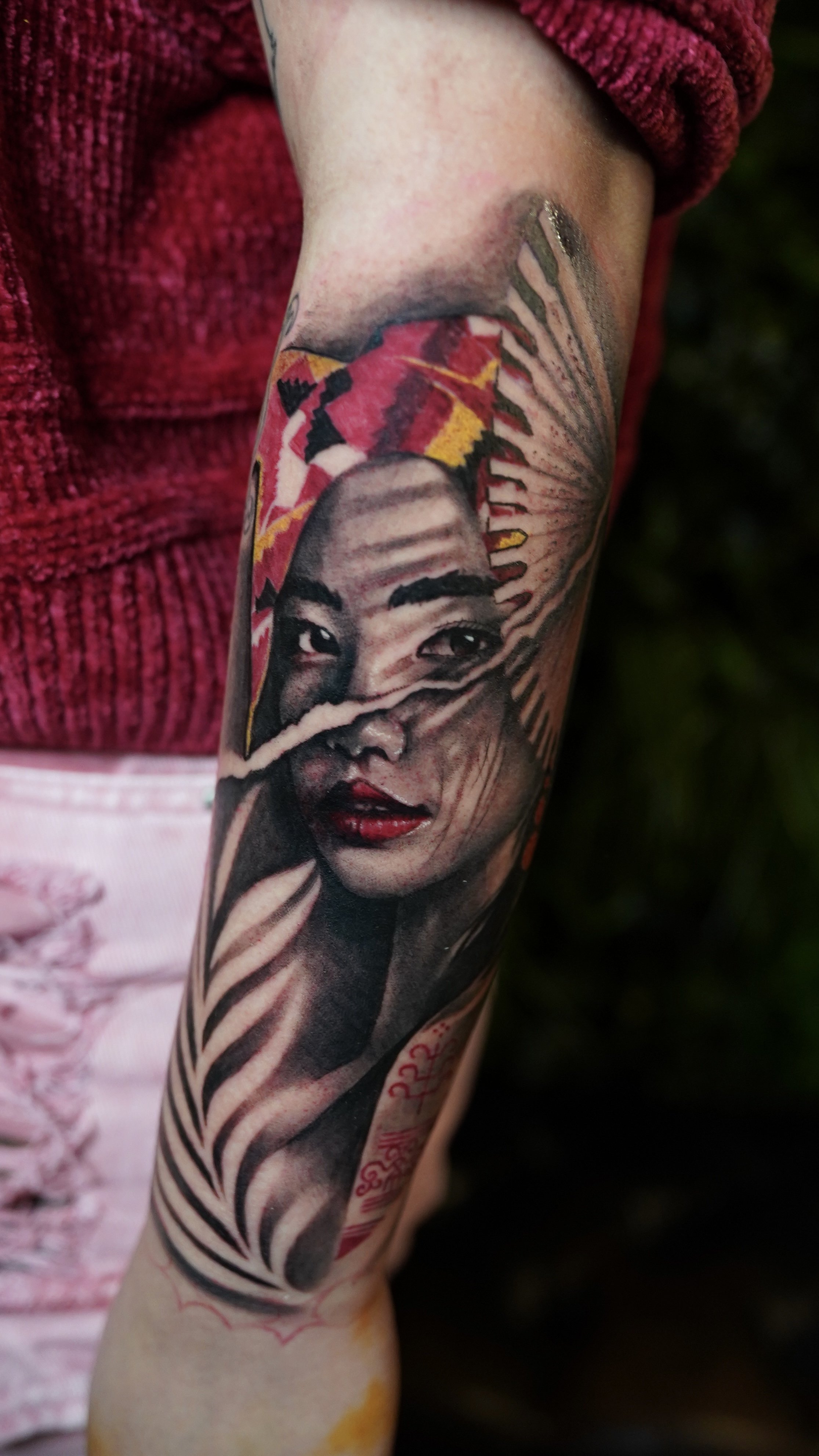 Close-up of a tattooed arm featuring a portrait of a woman with dark hair, red lipstick, and Asian features, with aesthetic elements surrounding her face.