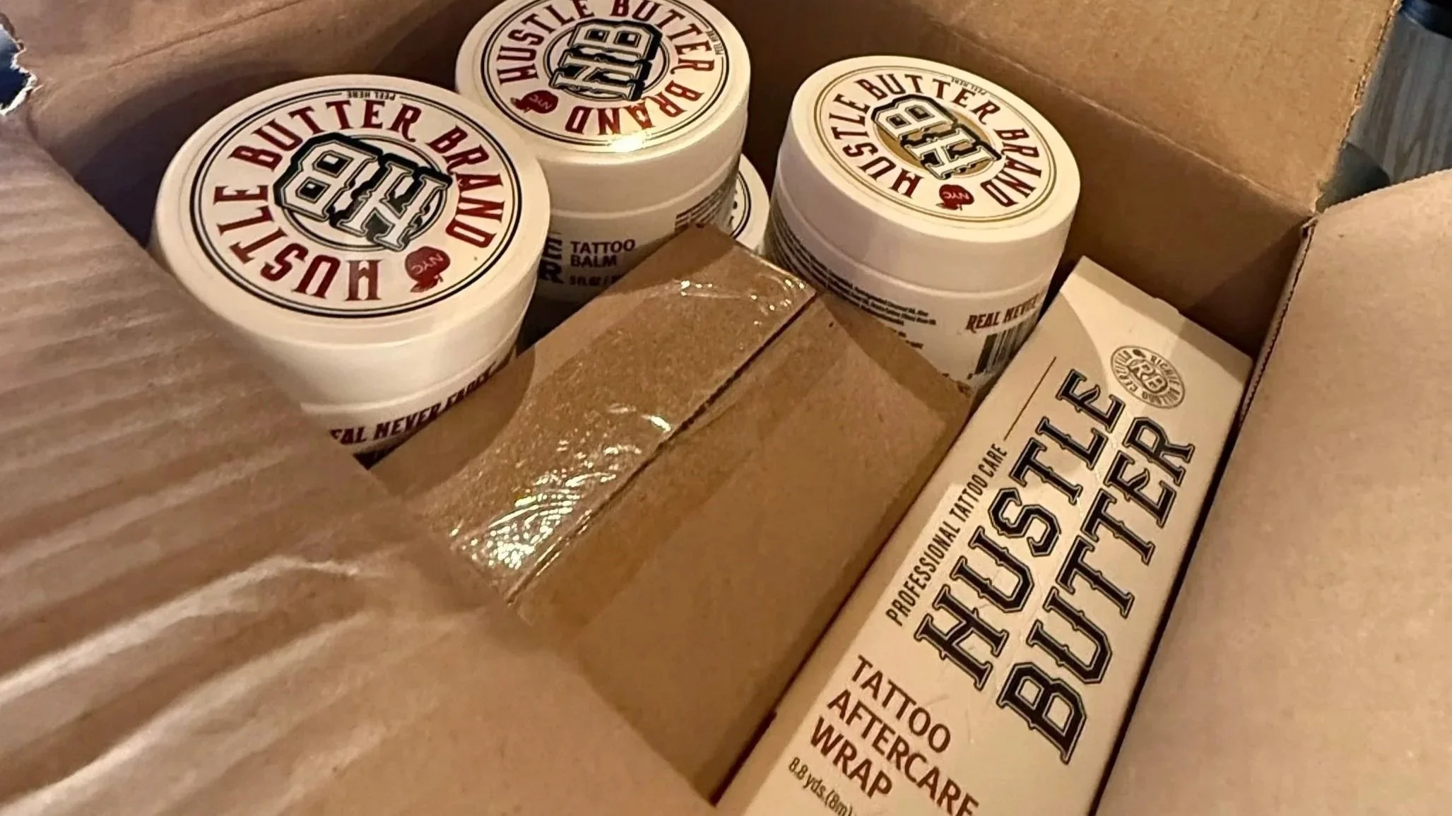 Several containers of Hustle Butter Deluxe tattoo aftercare balm inside a cardboard box.