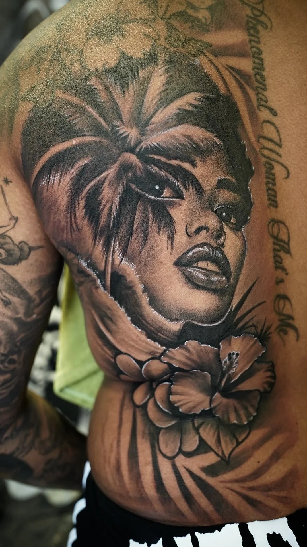 A detailed black and gray tattoo of a woman's face with prominent lips and eyes, surrounded by flowers and text on the person's upper arm and shoulder.