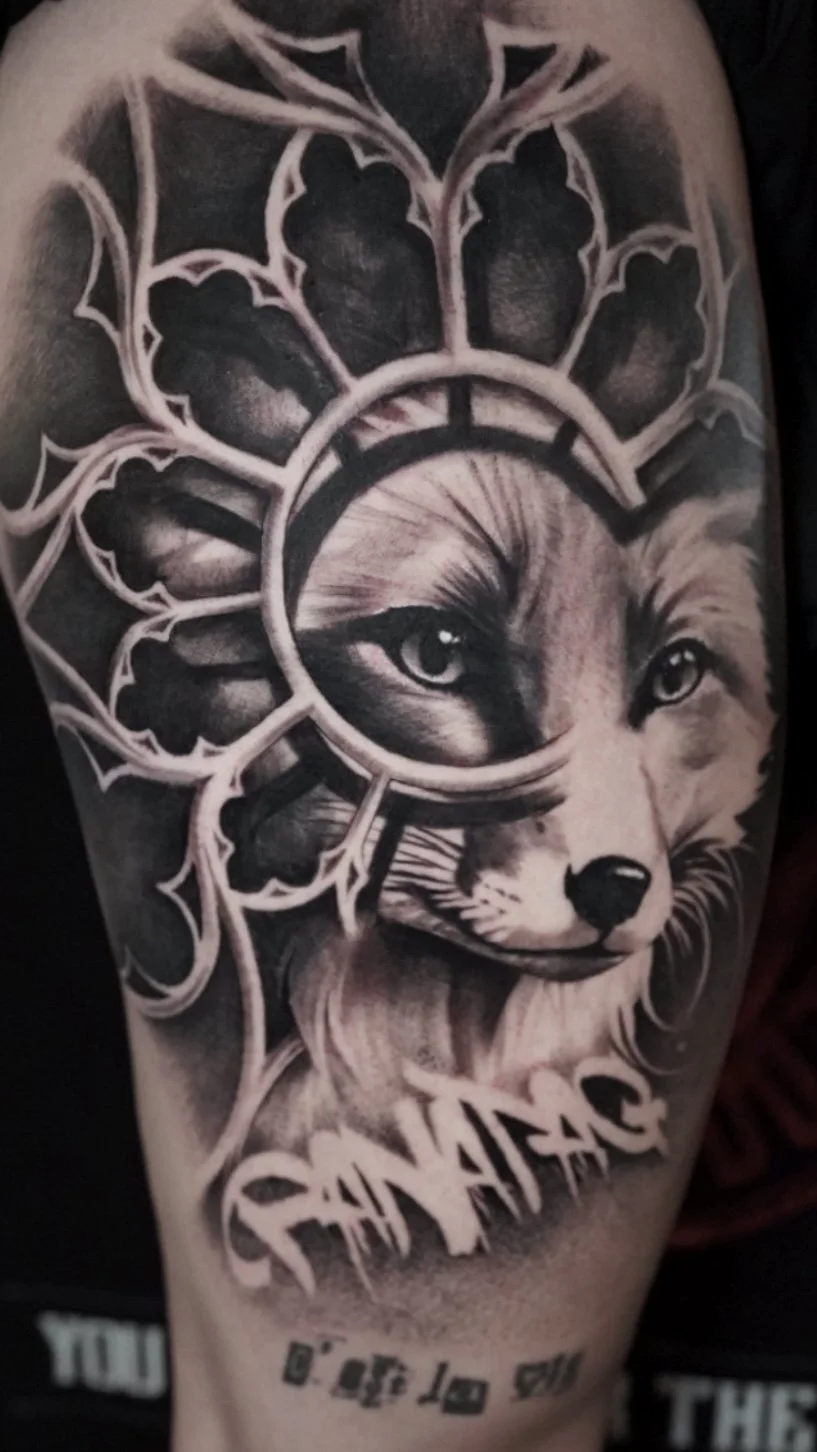 A black and gray tattoo of a wolf's face surrounded by stylized leaves and a circular frame, with the name 'Chamadra' written beneath in bold, artistic lettering.