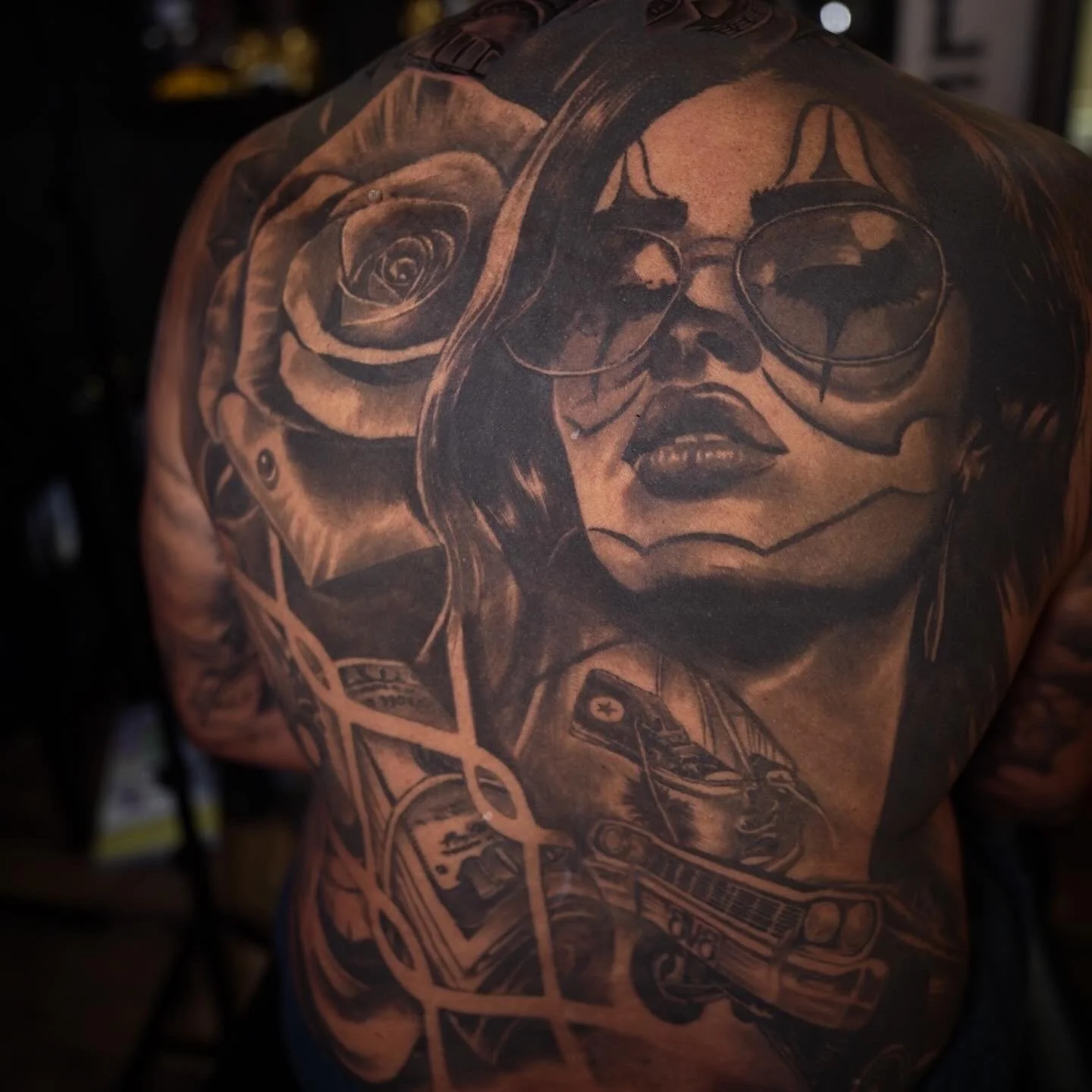 Black and gray tattoo of a woman's face wearing sunglasses, surrounded by roses and various elements including a motorcycle on a person's shoulder and upper arm.