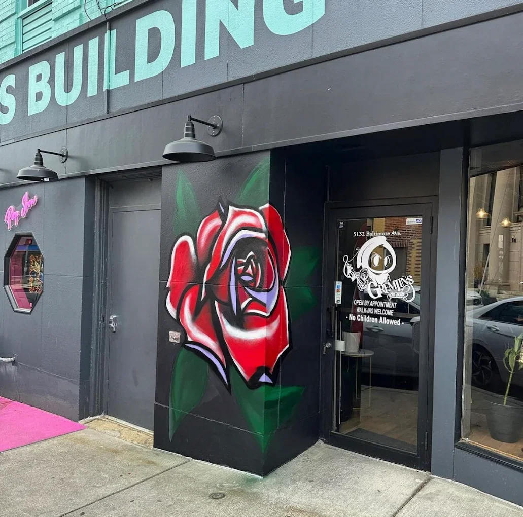 Store entrance with a large mural of a red rose painted on a black wall. The entrance door has a sign with the address 5132 Baltimore Ave., and text indicating the business, 'GEMINI & GEMS,' is open by appointment, welcomes walk-ins, and does not allow children.