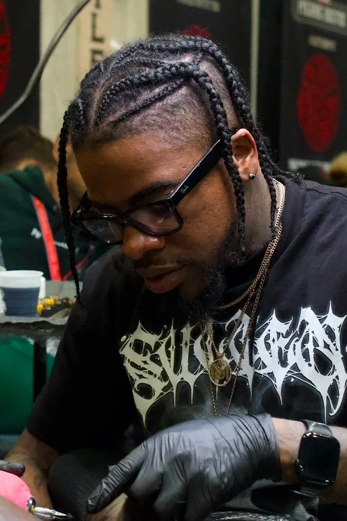 A man with braided hair, wearing glasses and black gloves, is focused on a task. He has earrings, layered necklaces, and a black graphic T-shirt, with a tattoo on his left arm. The background shows a busy setting with people and various items.