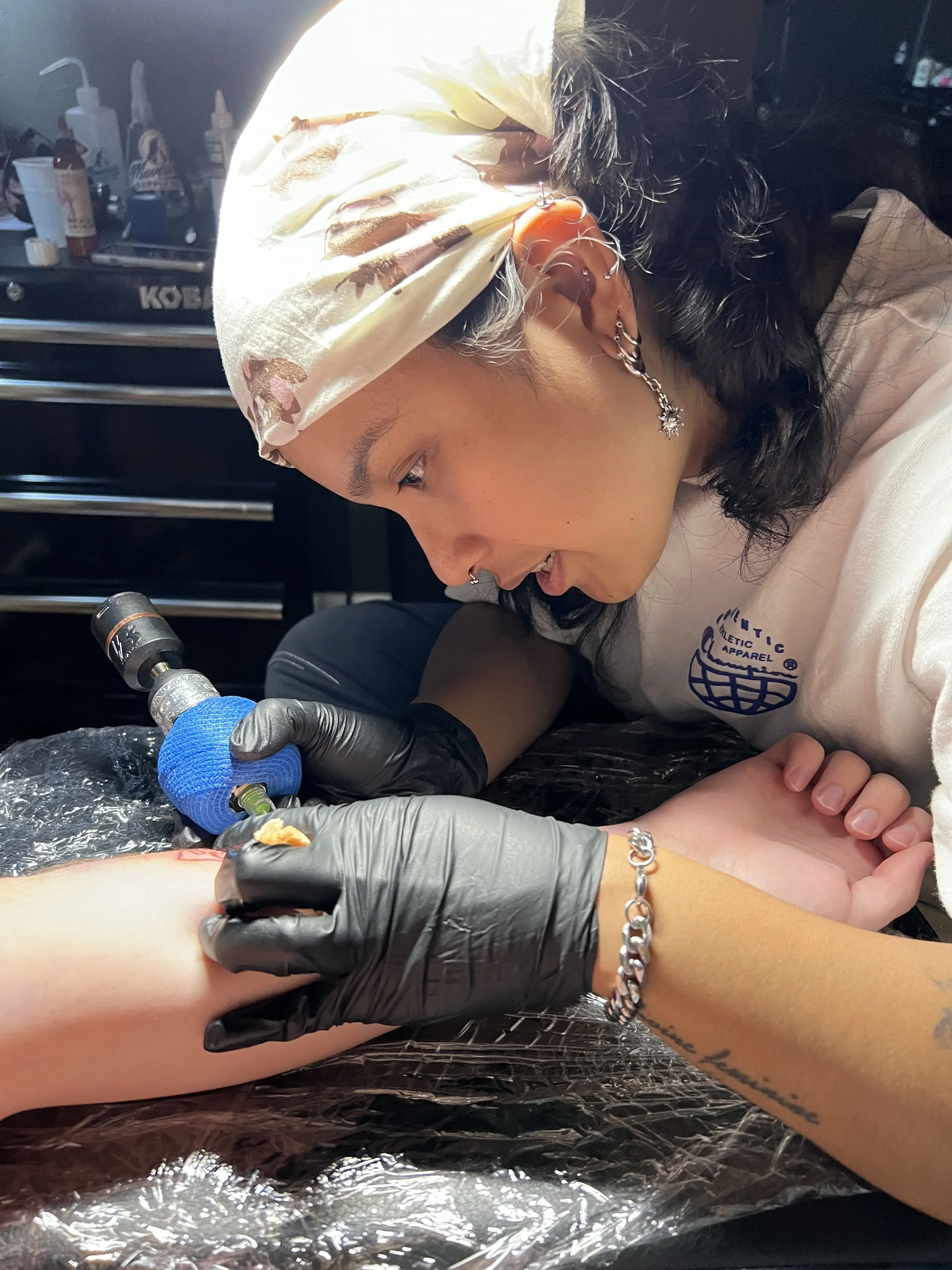 A person with dark hair, multiple earrings, and a floral scarf on their head is tattooing someone's arm. The person tattooing is wearing a silver bracelet and a glove on one hand. In the background, tattoo equipment and supplies are visible.