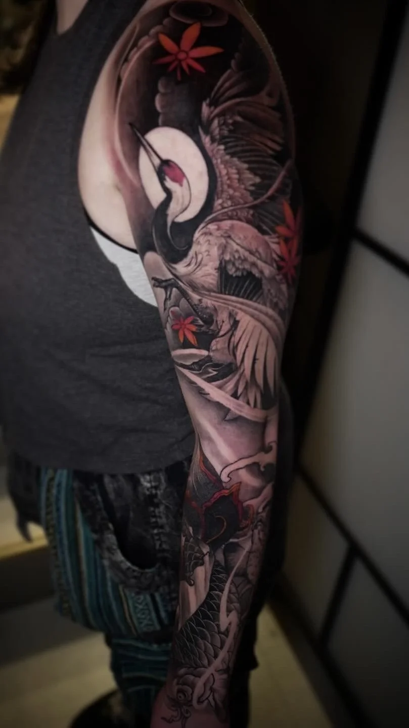 A detailed black and gray tattoo of a crane with red accents on the arm, featuring flowers and intricate patterns.