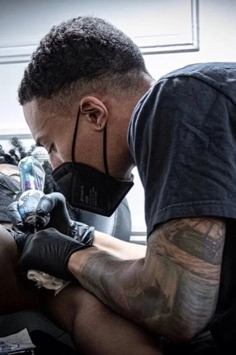 A tattooed man wearing a black face mask and gloves is tattooing someone's arm in a tattoo studio.