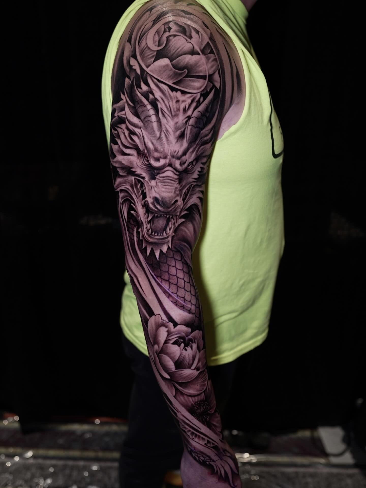 Close-up of a person's arm with a detailed black and gray tattoo of a fierce tiger, wearing a light yellow tank top, against a dark background.