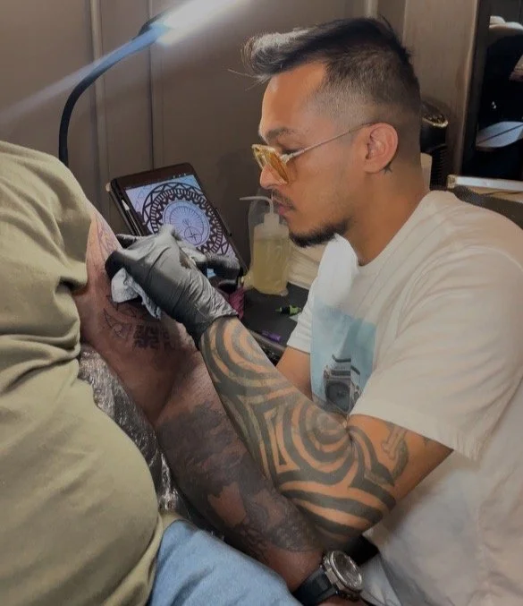 Tattoo artist tattooing a client's arm at a tattoo studio, with a tattoo design displayed on a tablet nearby, and a lamp illuminating the workspace.