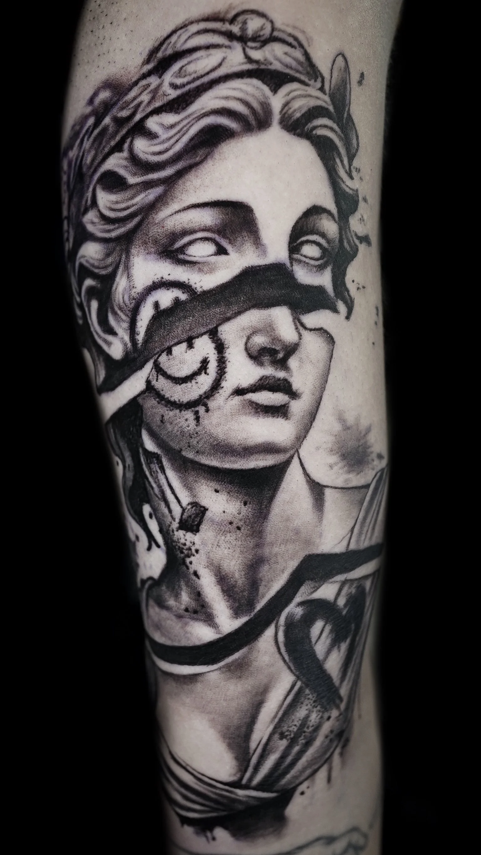 Black and gray tattoo of a woman's face with a blindfold, detailed hair, and decorative elements, covering a portion of an arm.