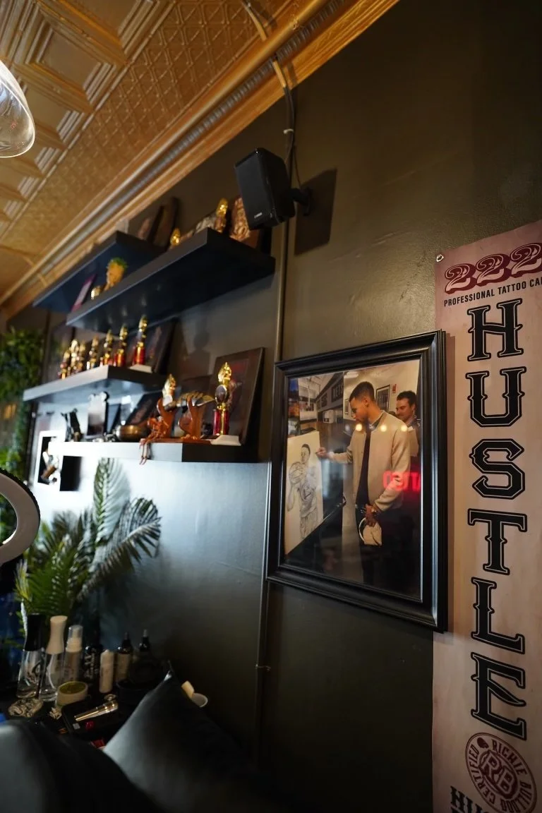Interior of a tattoo studio with framed artwork, a black shelf with trophies, and a leaning towards the event area that is decorated with plants and tattoo supplies.