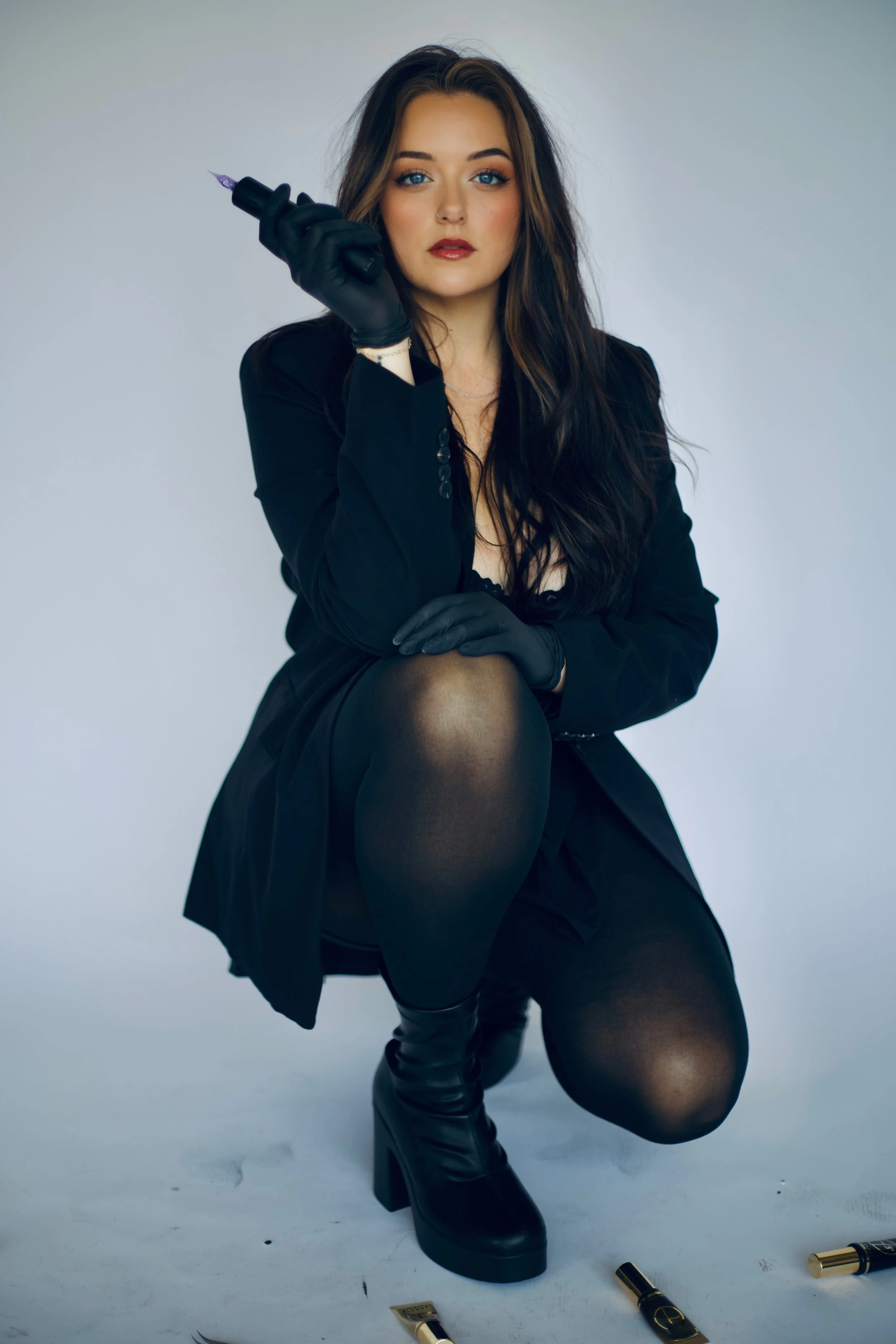 A young woman with long dark hair and blue eyes, dressed in black, holds a tattoo machine in her right hand. She is sitting with her right knee up, wearing black tights and thigh-high boots, against a plain light-colored background. There are makeup tubes scattered on the floor around her.