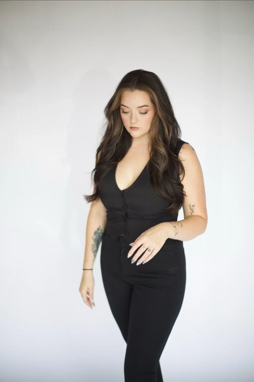 A woman with long brown hair wearing a black sleeveless top and black pants standing against a plain white background.