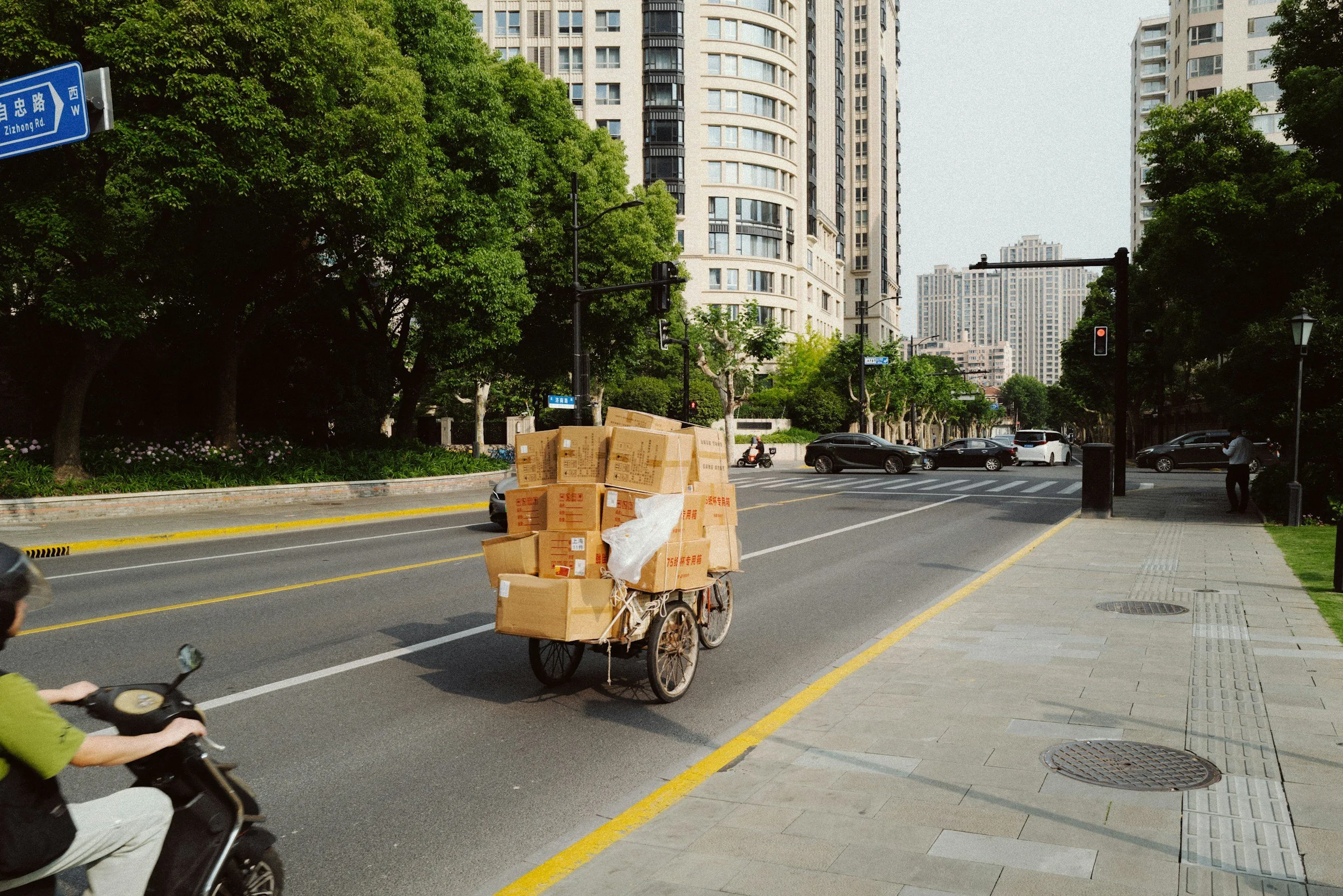A city street scene with high-rise buildings, green trees, parked cars, a traffic signal, and a person riding a scooter. A bicycle cart loaded with cardboard boxes is visible near the curb.