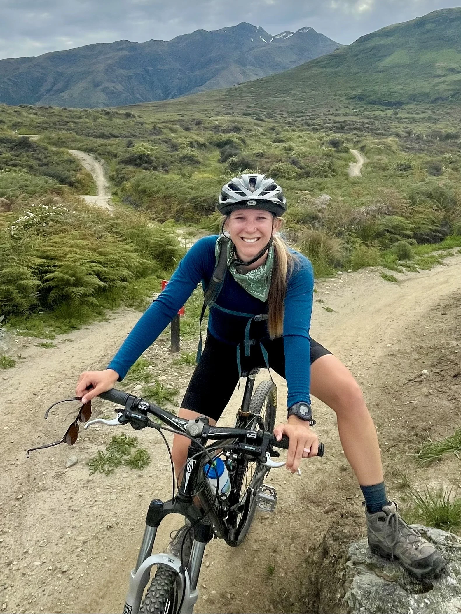 A woman smiling and wearing a helmet while riding a mountain bike on a dirt trail in a scenic outdoor landscape with mountains and green hills in the background.