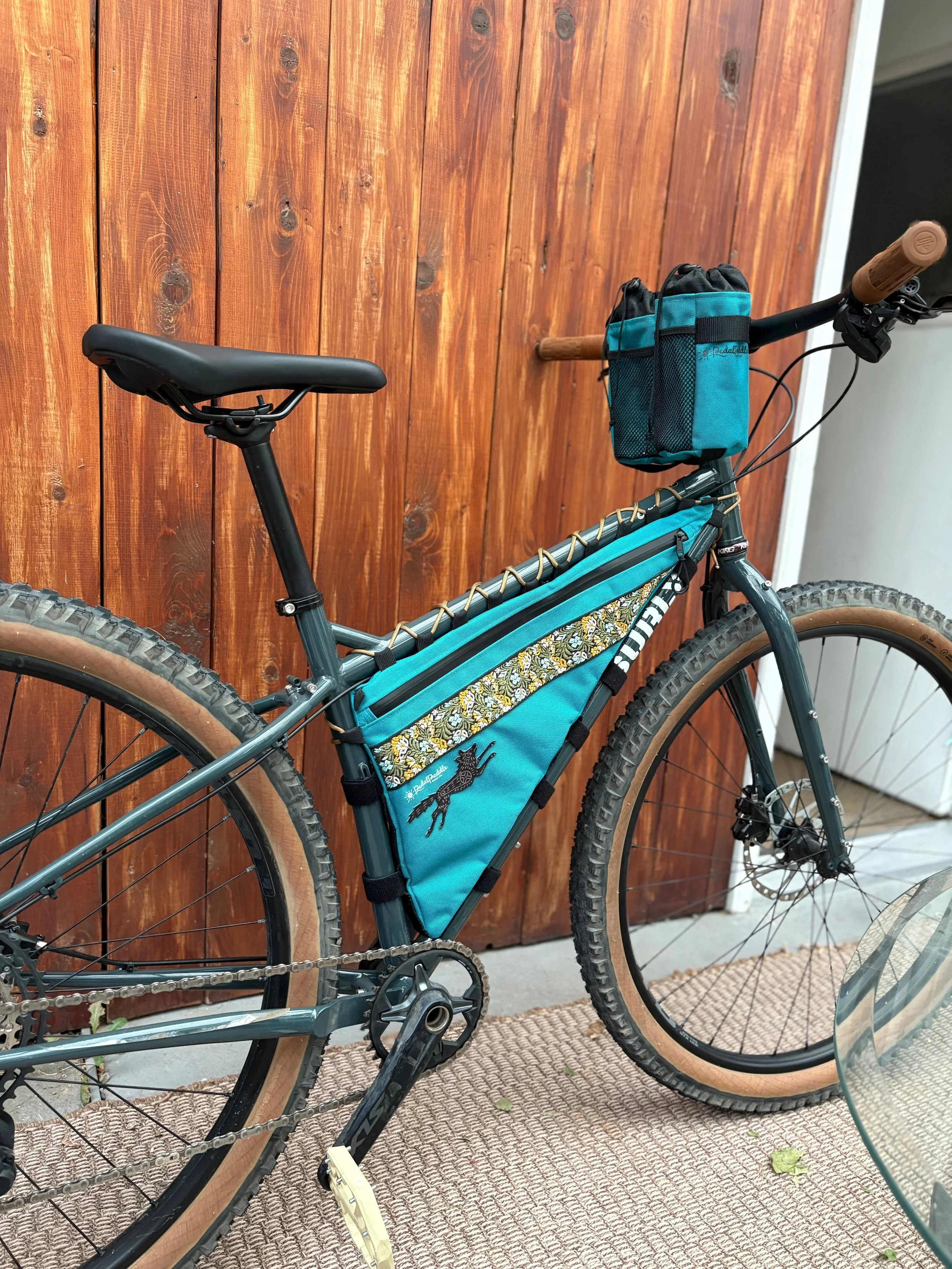A womens gravel bike with a turquoise frame bag, black seat, and brown tires, parked next to a wooden fence; two stem bags are attached to the handlebars