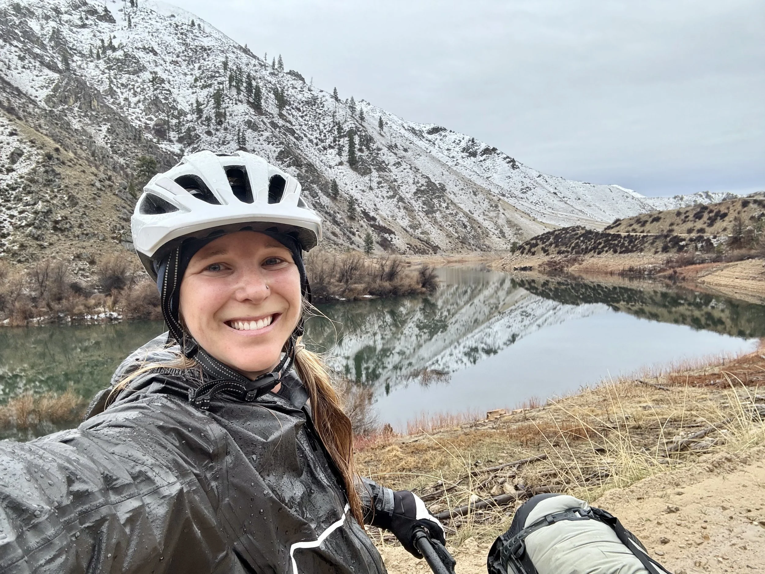 A smiling woman wearing a bike helmet and rain jacket taking a selfie near a river, with snow-covered mountains and trees in the background on a cloudy day.