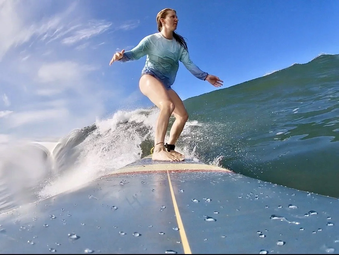 A woman surfing on a wave in the ocean, wearing a long-sleeve shirt and shorts, with the sky in the background.