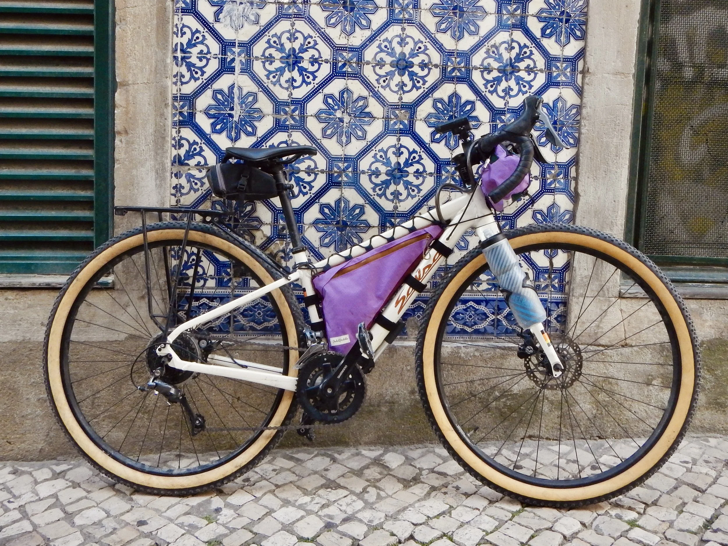A white bicycle parked against a wall with decorative blue and white ceramic tiles and two windows, one with green shutters and the other with a metal grate, on a cobblestone street.