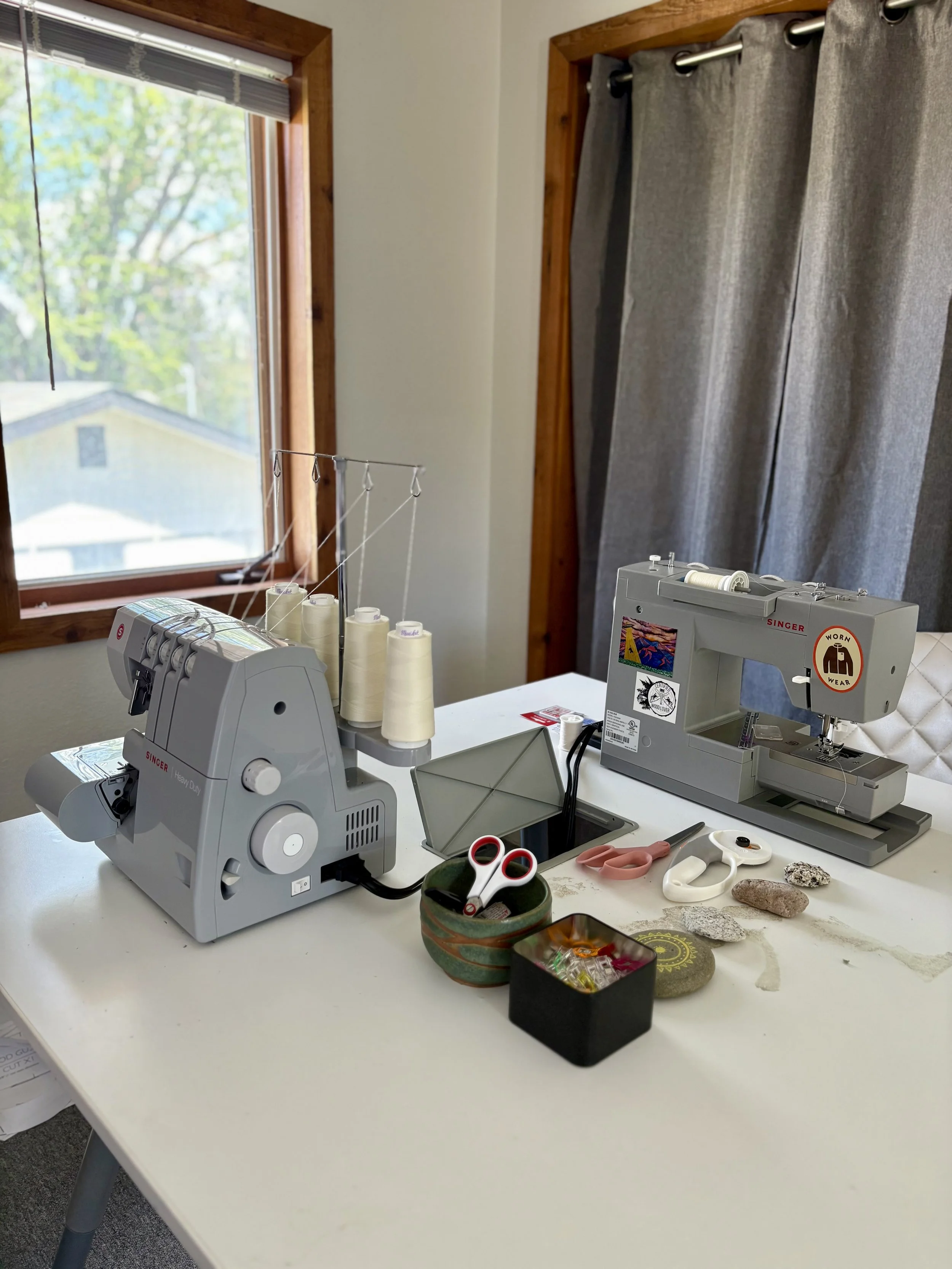 A sewing workspace with two sewing machines on a white table, scissors, fabric, and sewing accessories; window with green trees outside and gray curtains.