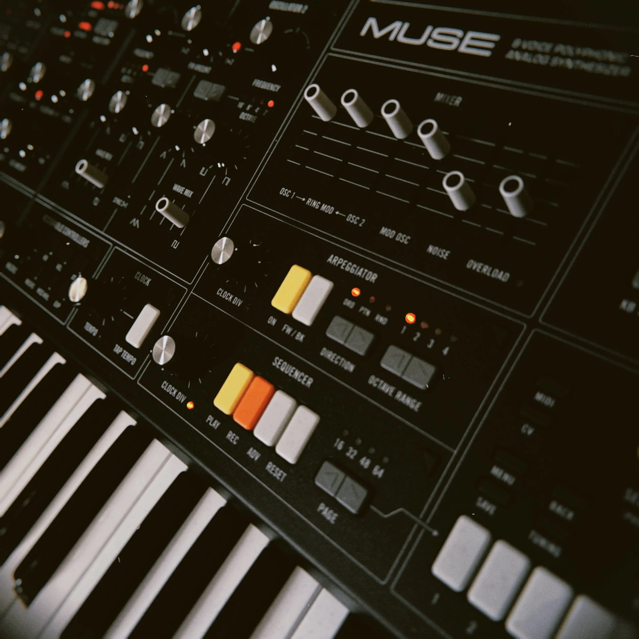 Close-up of a DJ or music producer's equipment, featuring an analog synthesizer or drum machine with knobs, sliders, and buttons, including orange, white, and yellow controls.