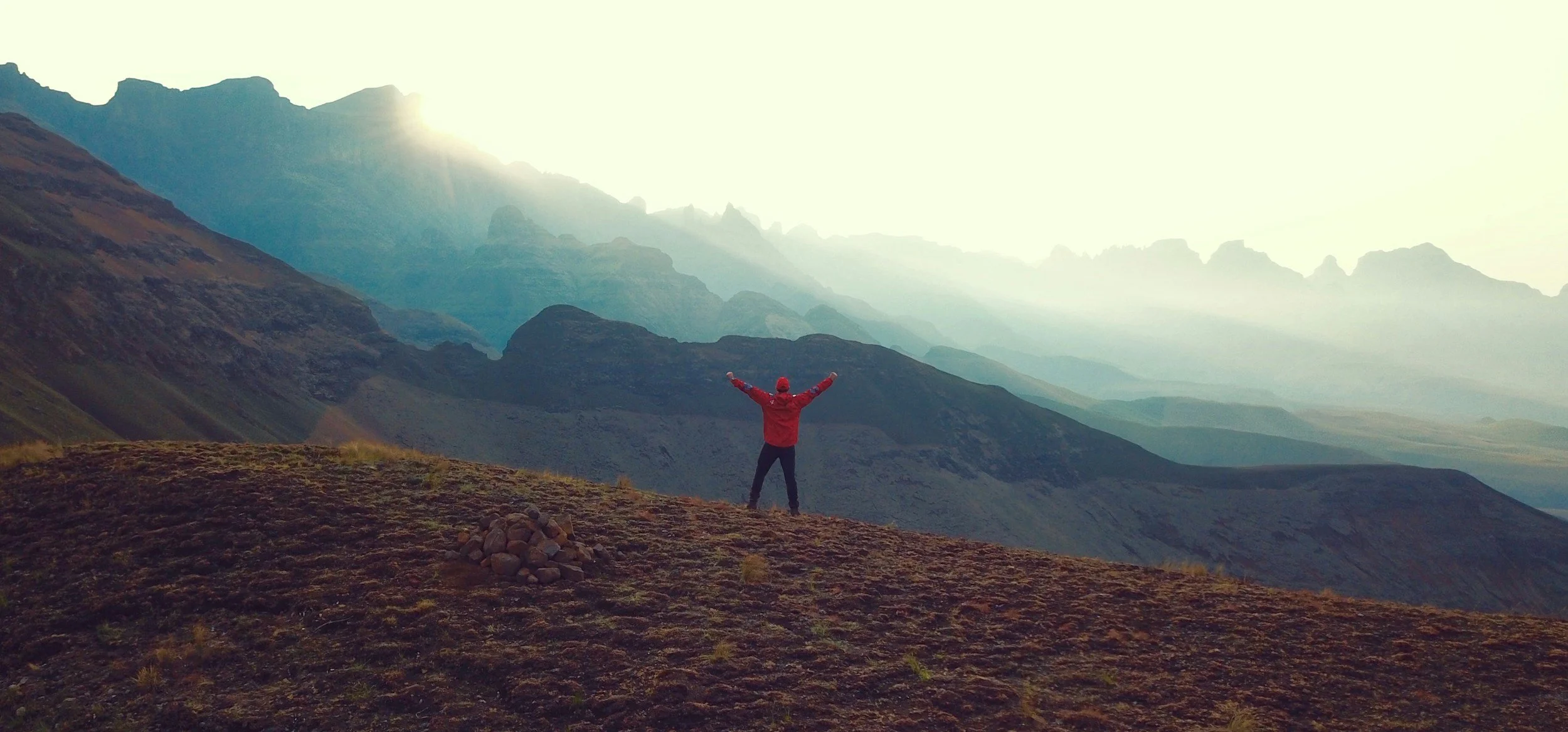 Person in red jacket and hat standing on a mountain with arms raised, overlooking a range of mountains bathed in sunlight. Feeling successful and proud.