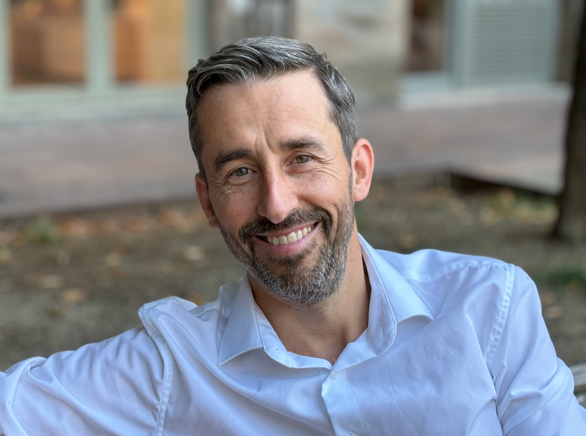 Portrait of a smiling man with short gray hair and a beard, wearing a white shirt, outdoors with blurred background.