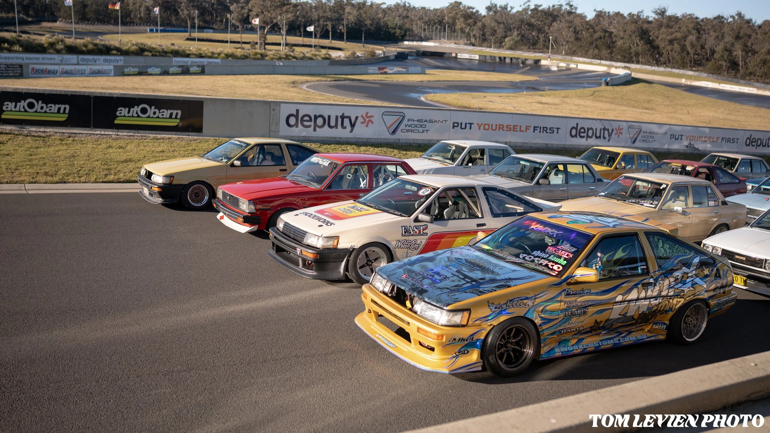 Multiple vintage race cars with colorful decals parked in a row at Pheasant Wood Circuit racetrack, with a view of the tracks and trees in the background.