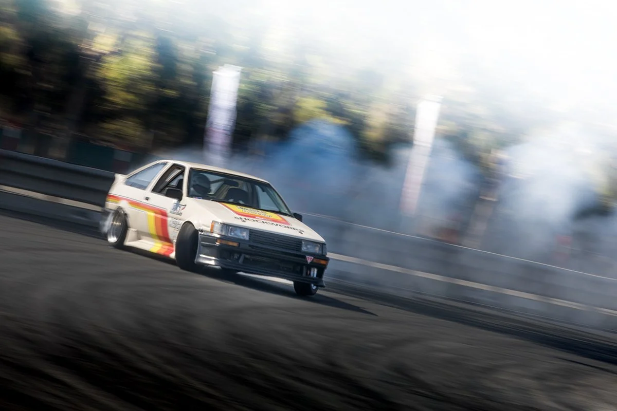 A white race car with colorful racing stripes drifting at high speed on a race track, producing smoke from the tires, with blurred background trees and trackside banners.