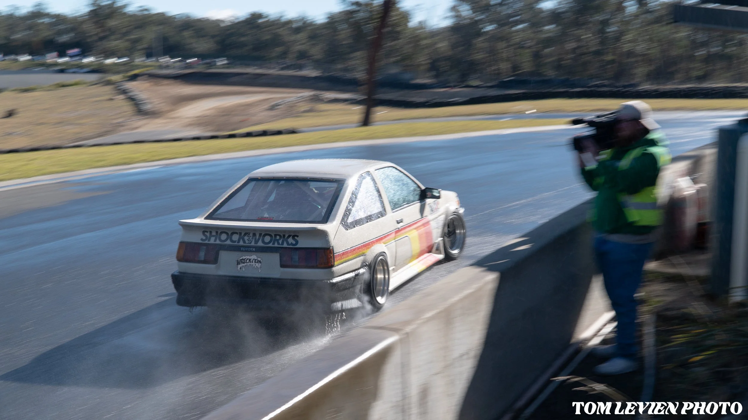 A race car on a wet track with water splashing behind it, and a camera operator recording from the side. The background shows a rural area with trees and a fence.