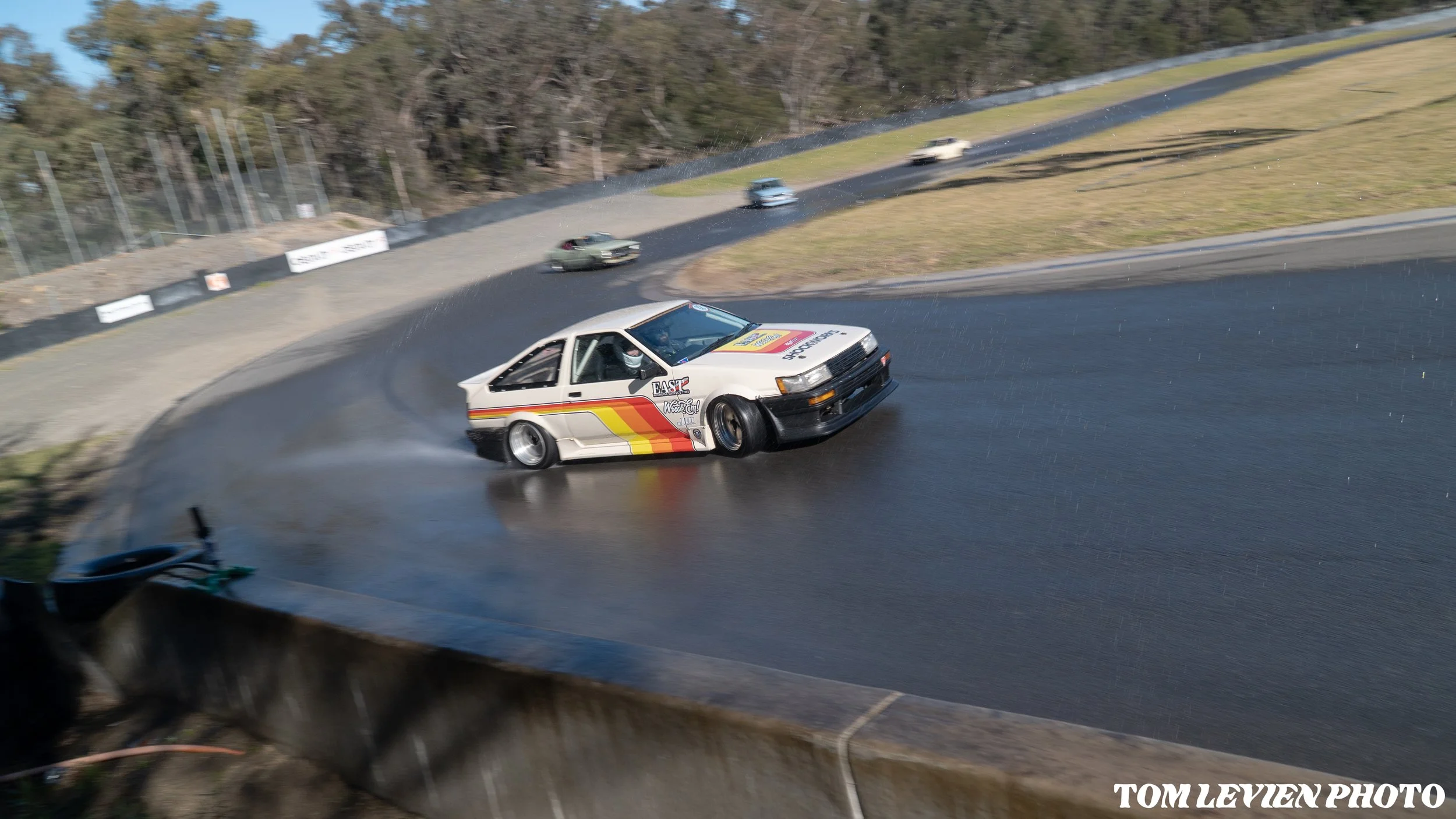 A white race car with red, yellow, and black stripes navigating a wet, curved race track with other cars behind. Trees line the background. The photo was taken from a lower vantage point near a barrier.