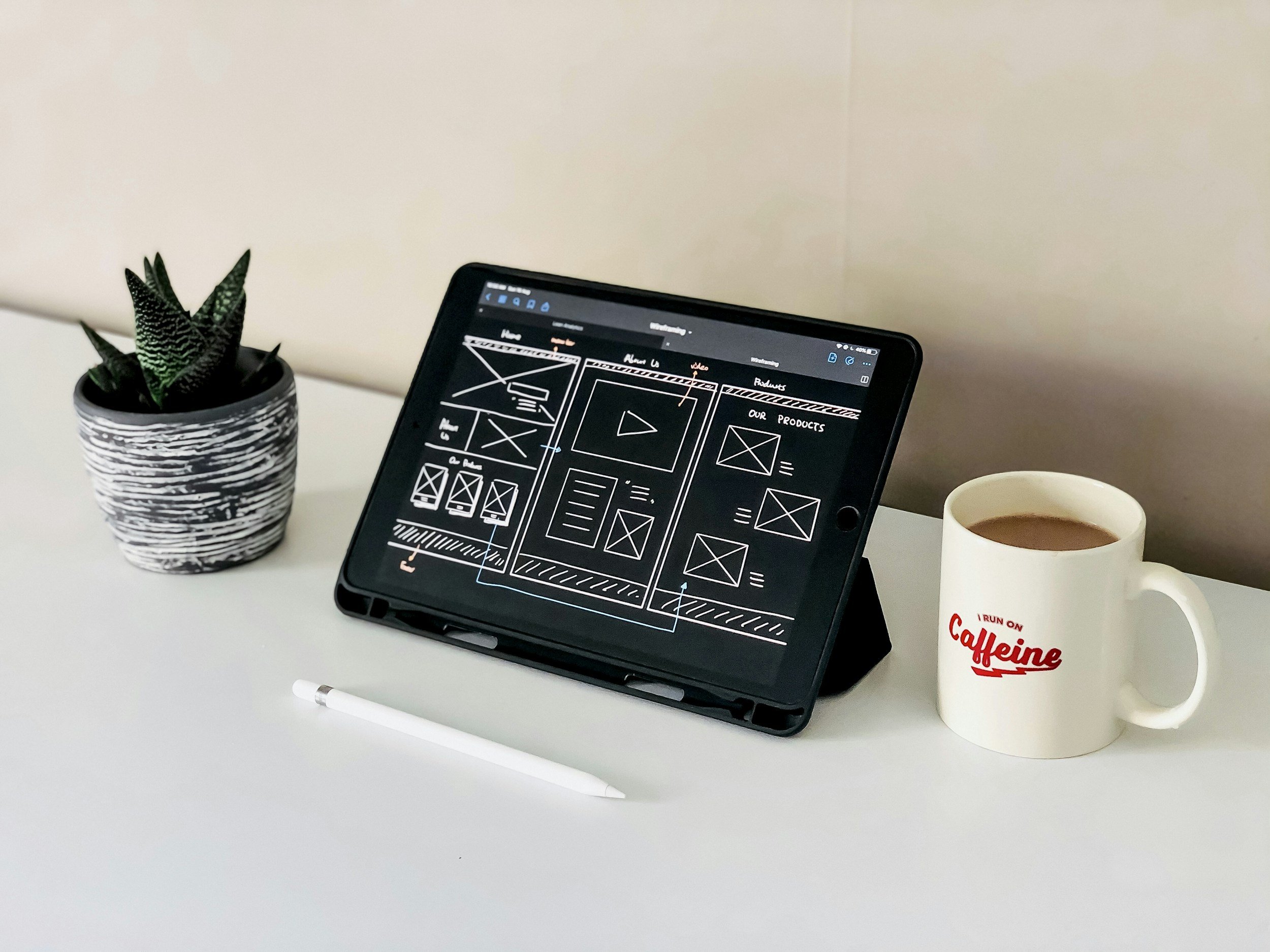 A white desk with a potted plant, a tablet displaying a wireframe design, a white stylus, a white mug with red text, and a cup of coffee.