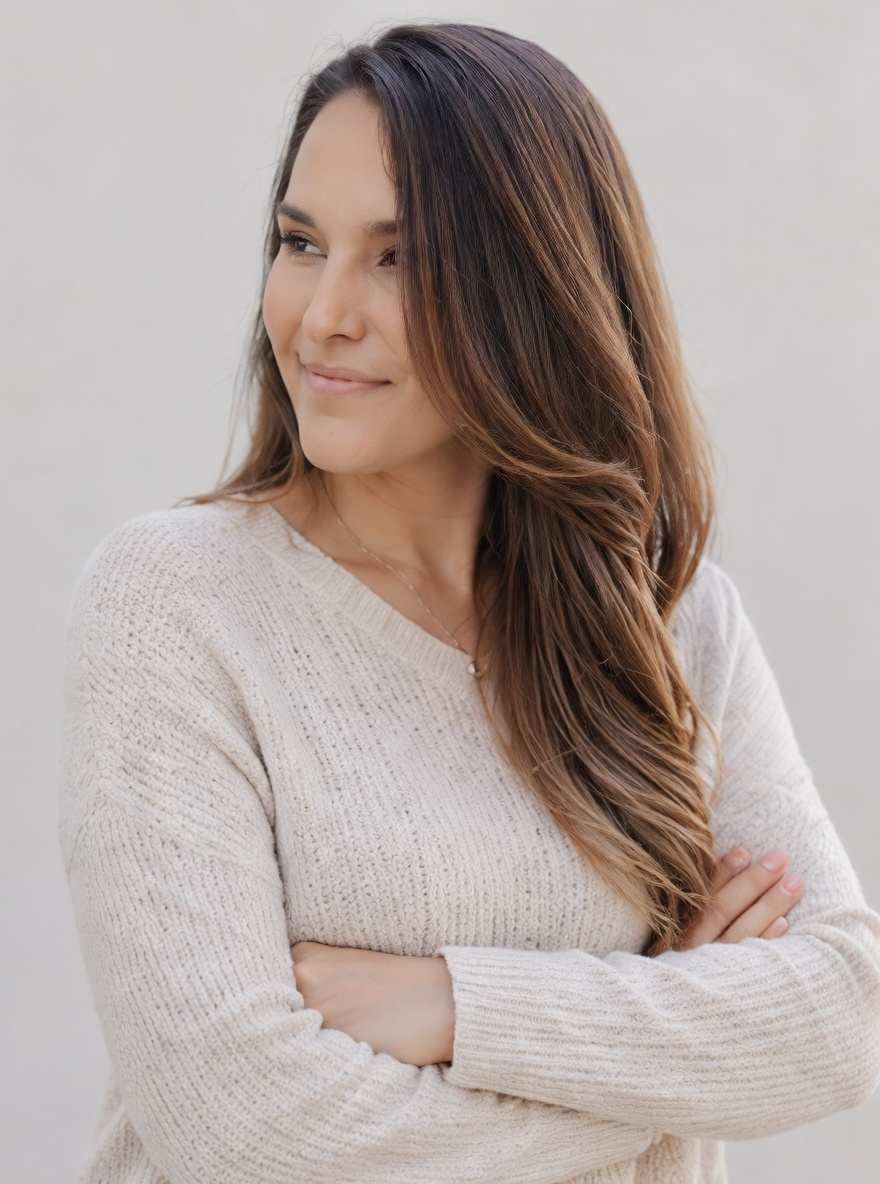 A woman with long, wavy brown hair wearing a cream knit sweater looks to her left with a slight smile, standing against a plain light gray background.