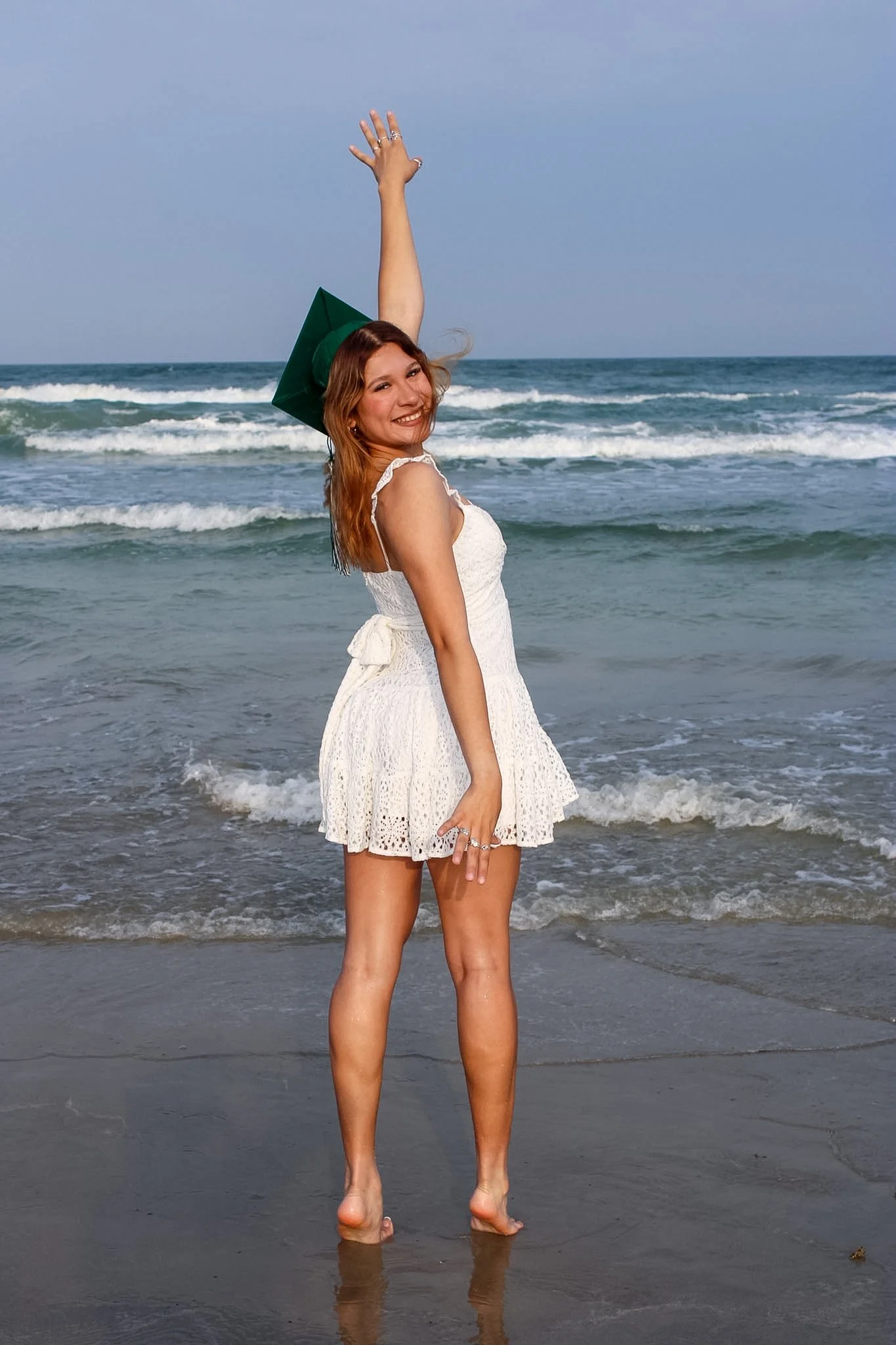 A smiling woman in a white dress with a green graduation cap, standing on wet sand at the beach with waves in the background, raising her right arm.