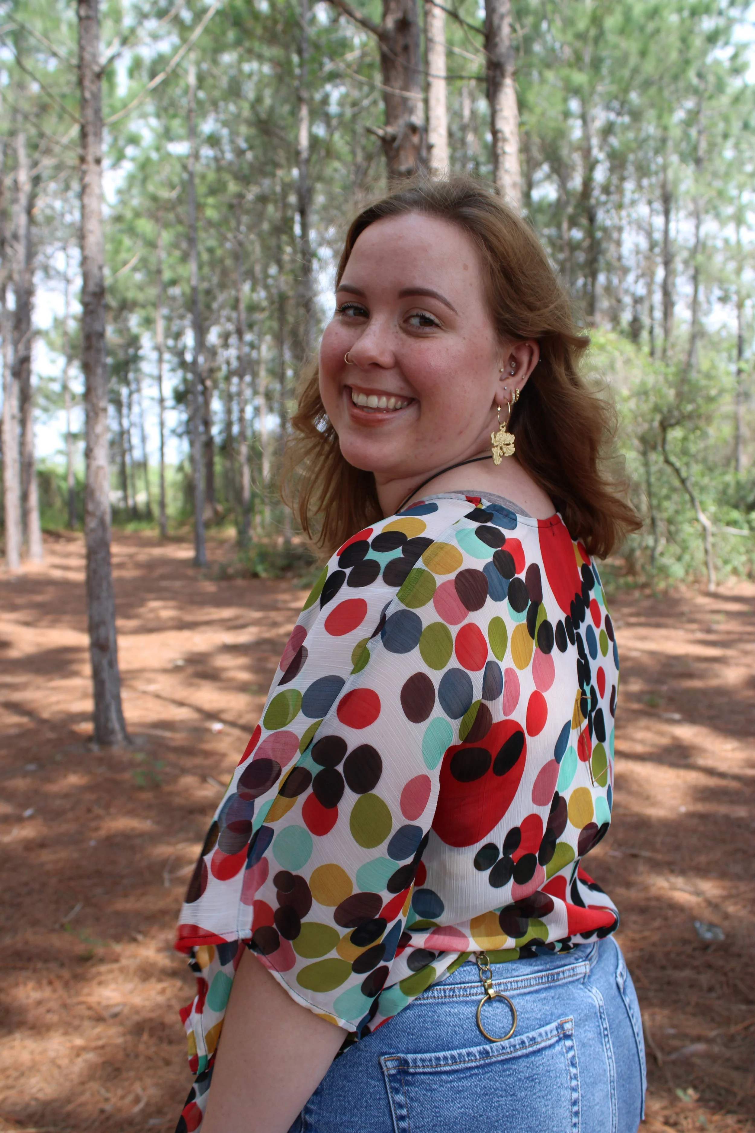 A young woman with red hair wearing a multicolored polka dot top and light blue jeans, smiling in an outdoor wooded area.