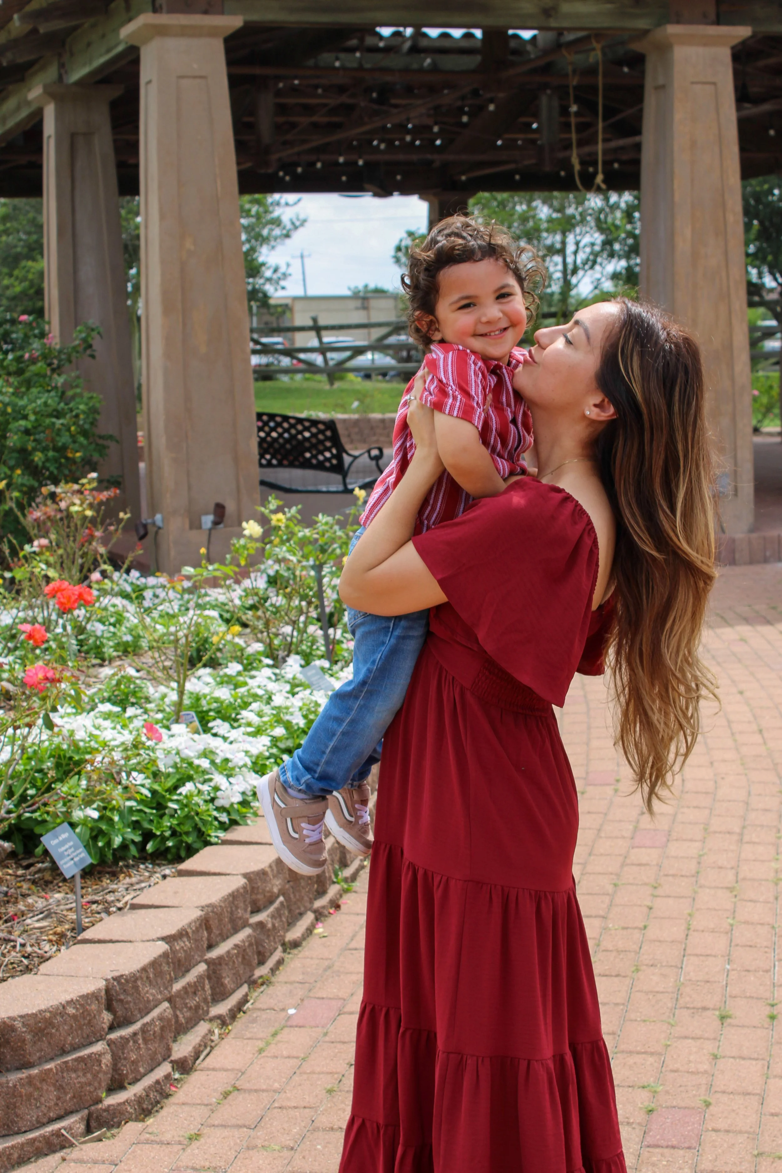 A woman with long, wavy hair, wearing a red dress, is holding a smiling young boy with curly hair, wearing a red and white striped shirt and jeans. They are outdoors in a garden with flowering plants and a wooden pergola in the background.