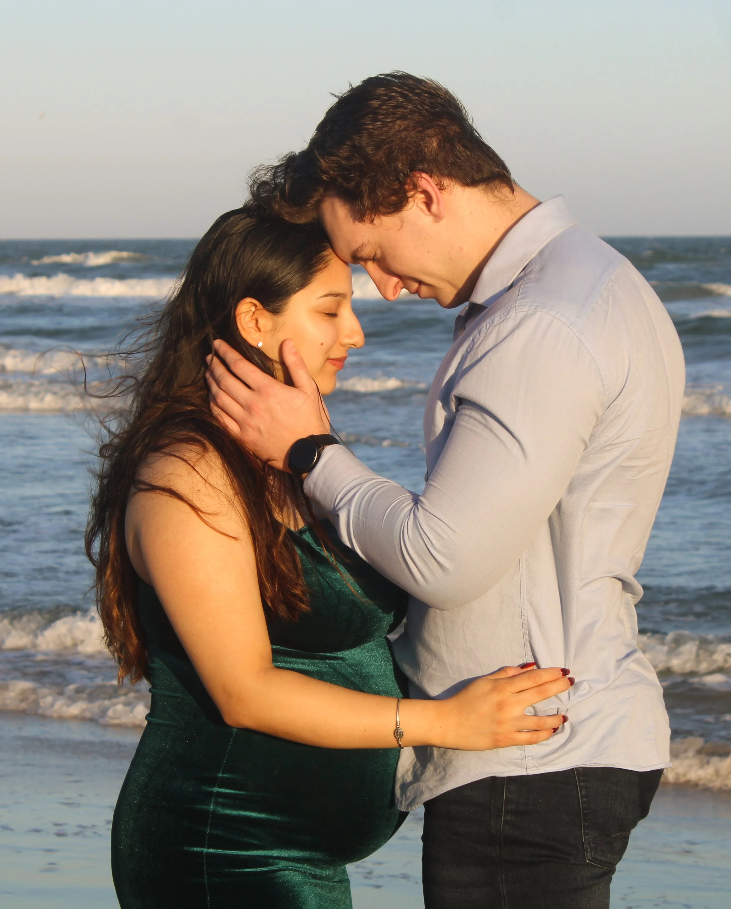 A couple stands close together on a beach, forehead to forehead, with eyes closed, holding each other affectionately during sunset.